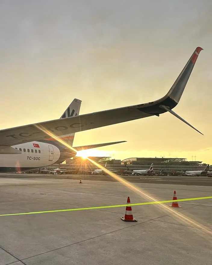 Flugzeugflügel mit Sonnenuntergang im Hintergrund auf einem Flughafenrollfeld mit weiteren Flugzeugen und Gebäuden im Hintergrund.