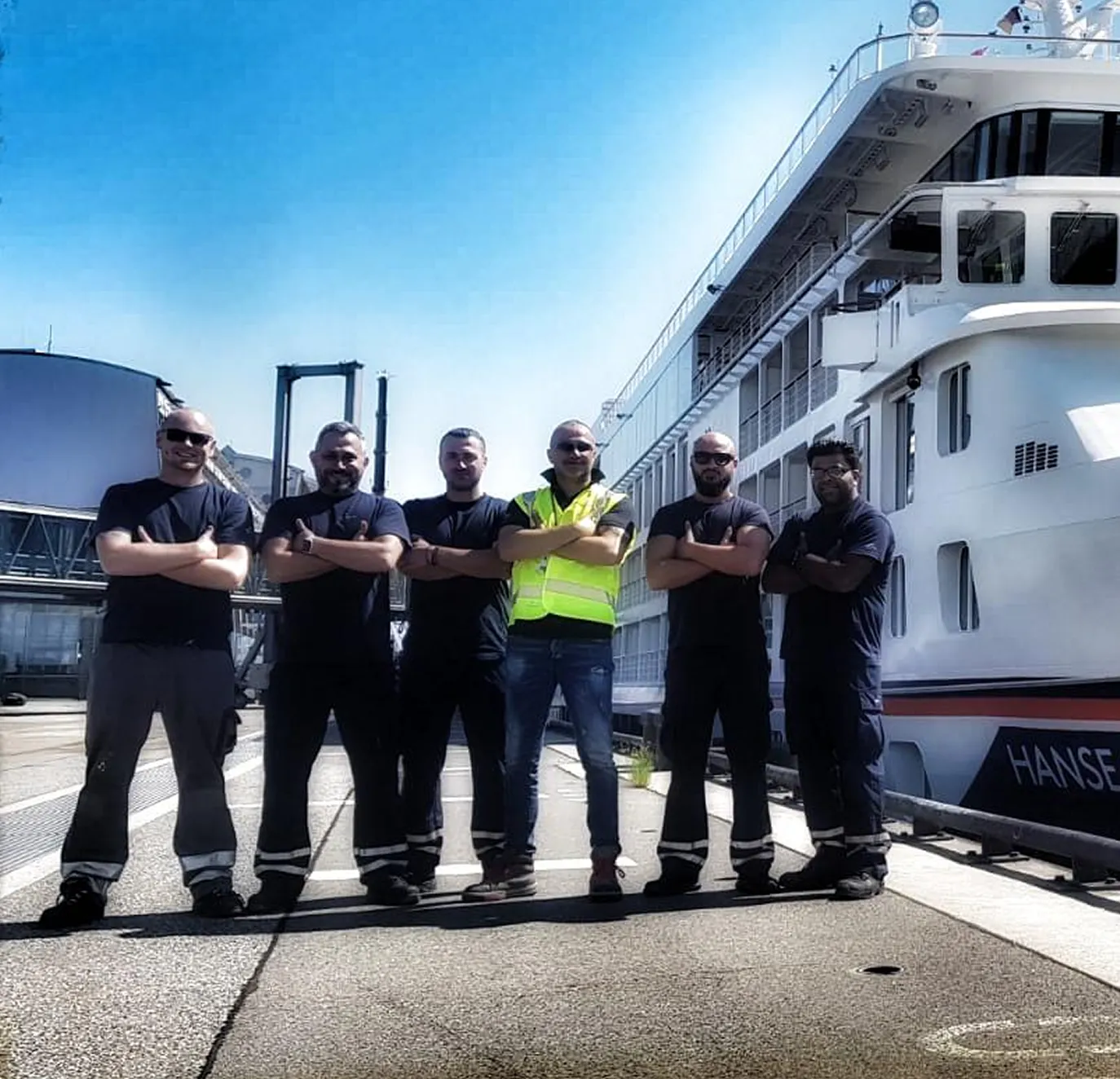 Sechs Männer mit verschränkten Armen stehen am Hafen vor einem großen weißen Schiff namens Hanse.