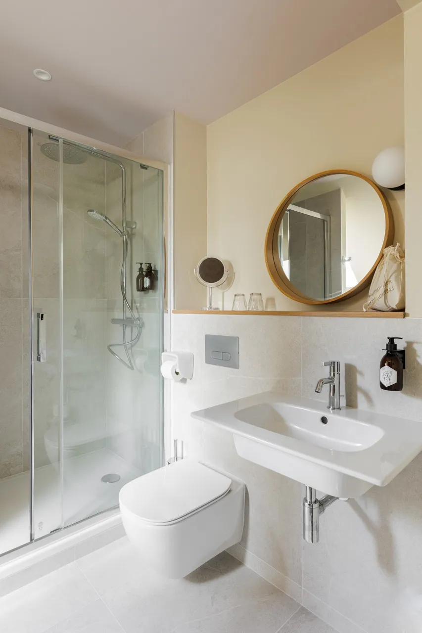 Salle de bain moderne avec une douche à l'italienne avec porte en verre, un lavabo blanc, un miroir rond en bois et des accessoires épurés.