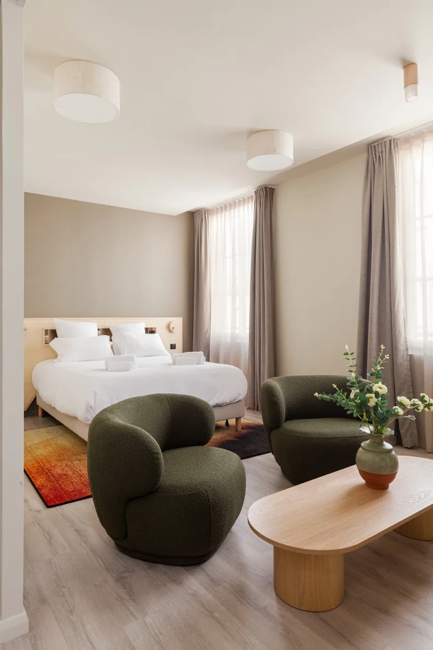 Chambre moderne avec un grand lit double, deux fauteuils verts et une table basse en bois avec un vase de fleurs.