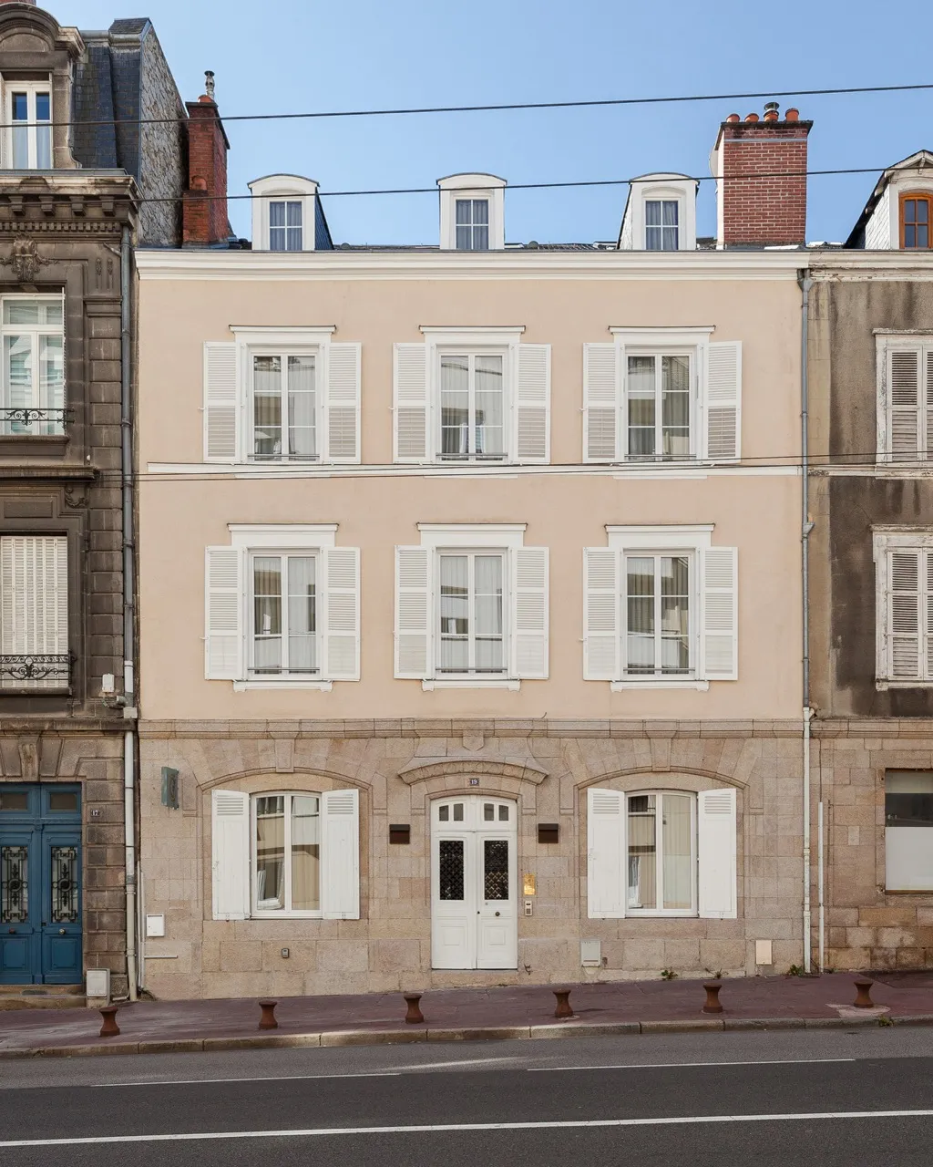 Façade d'un bâtiment à trois étages avec volets blancs ouverts sur les fenêtres, porte d'entrée blanche au centre et trottoir devant.