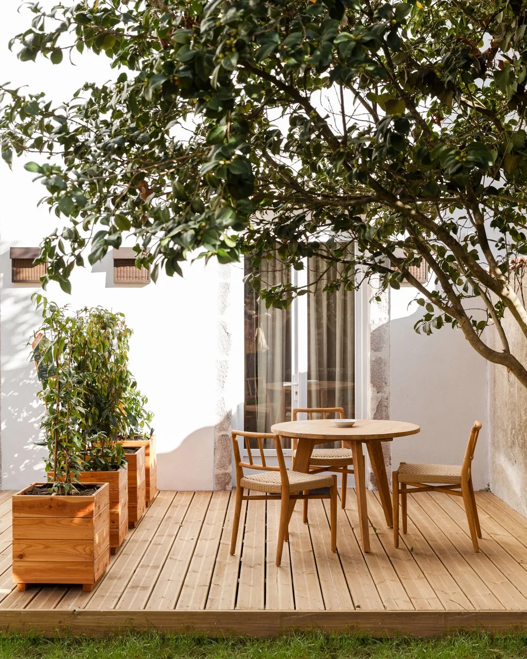 Terrasse en bois avec une table ronde et trois chaises en bois sous un arbre avec des plantes en pots.