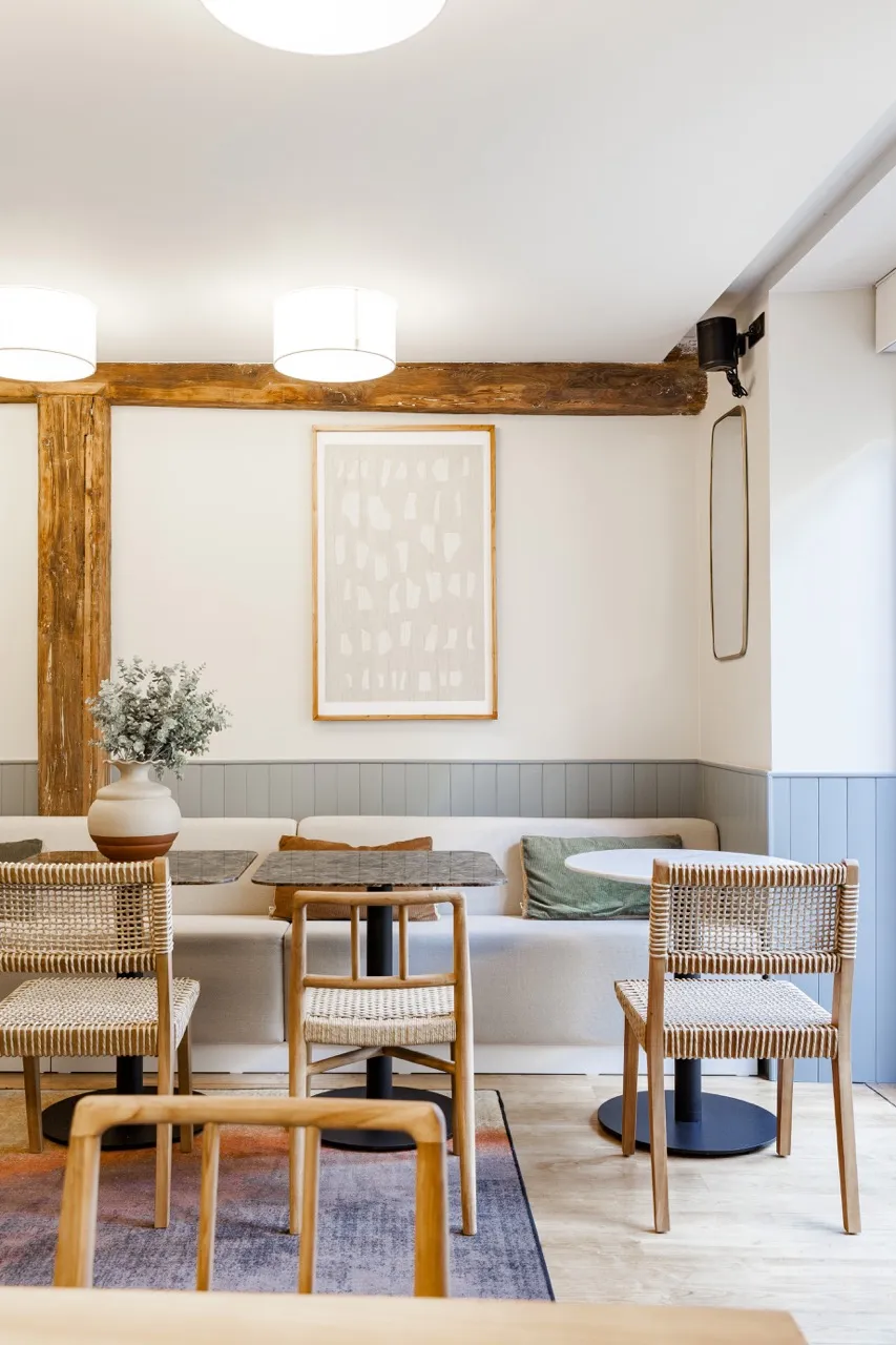 Intérieur d'un café avec tables, chaises en bois tressé, banquette blanche, coussins et décor minimaliste avec cadres et poutres apparentes.