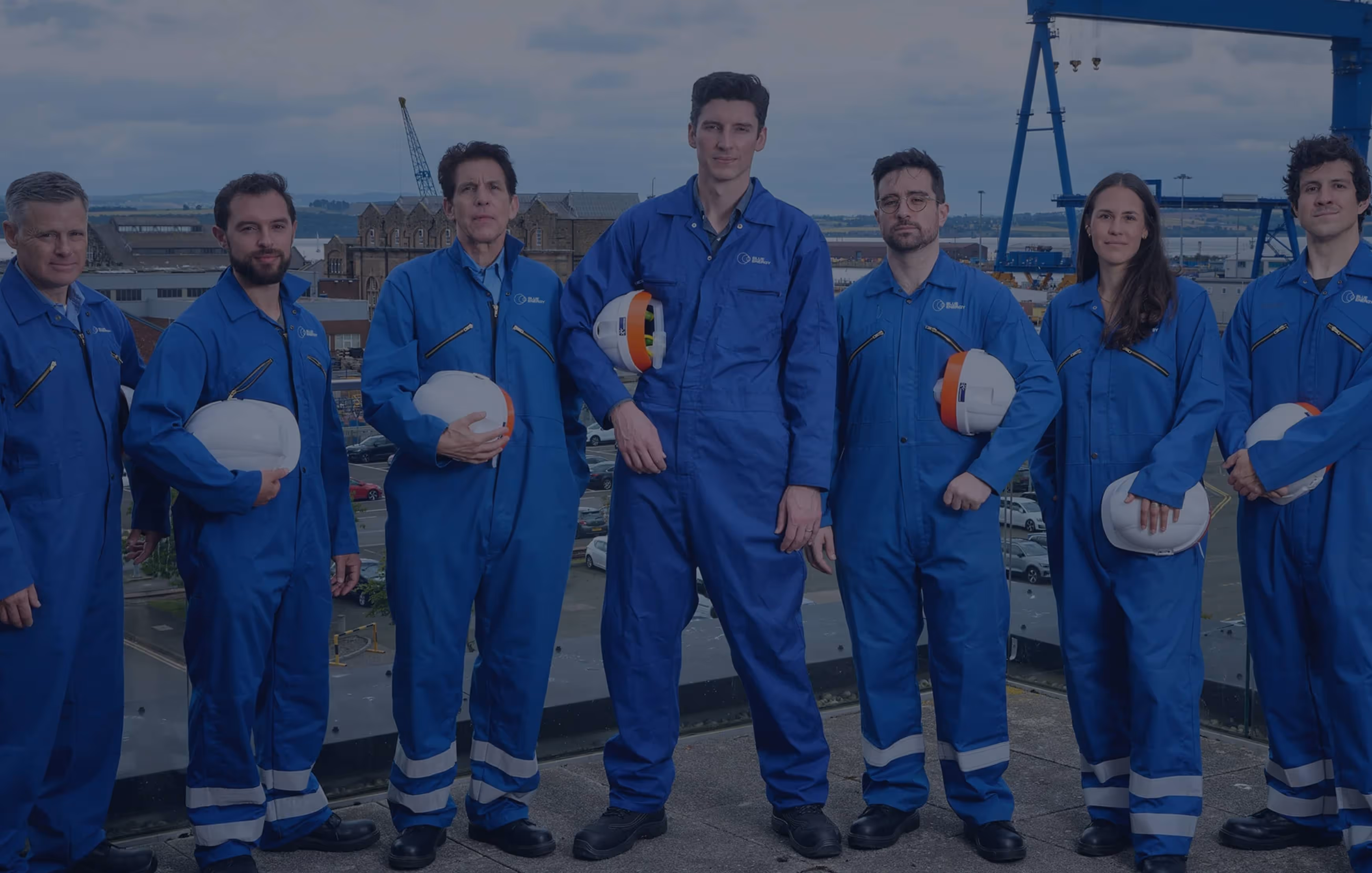 Group of seven engineers in blue coveralls holding white safety helmets on a rooftop with an industrial harbor background.