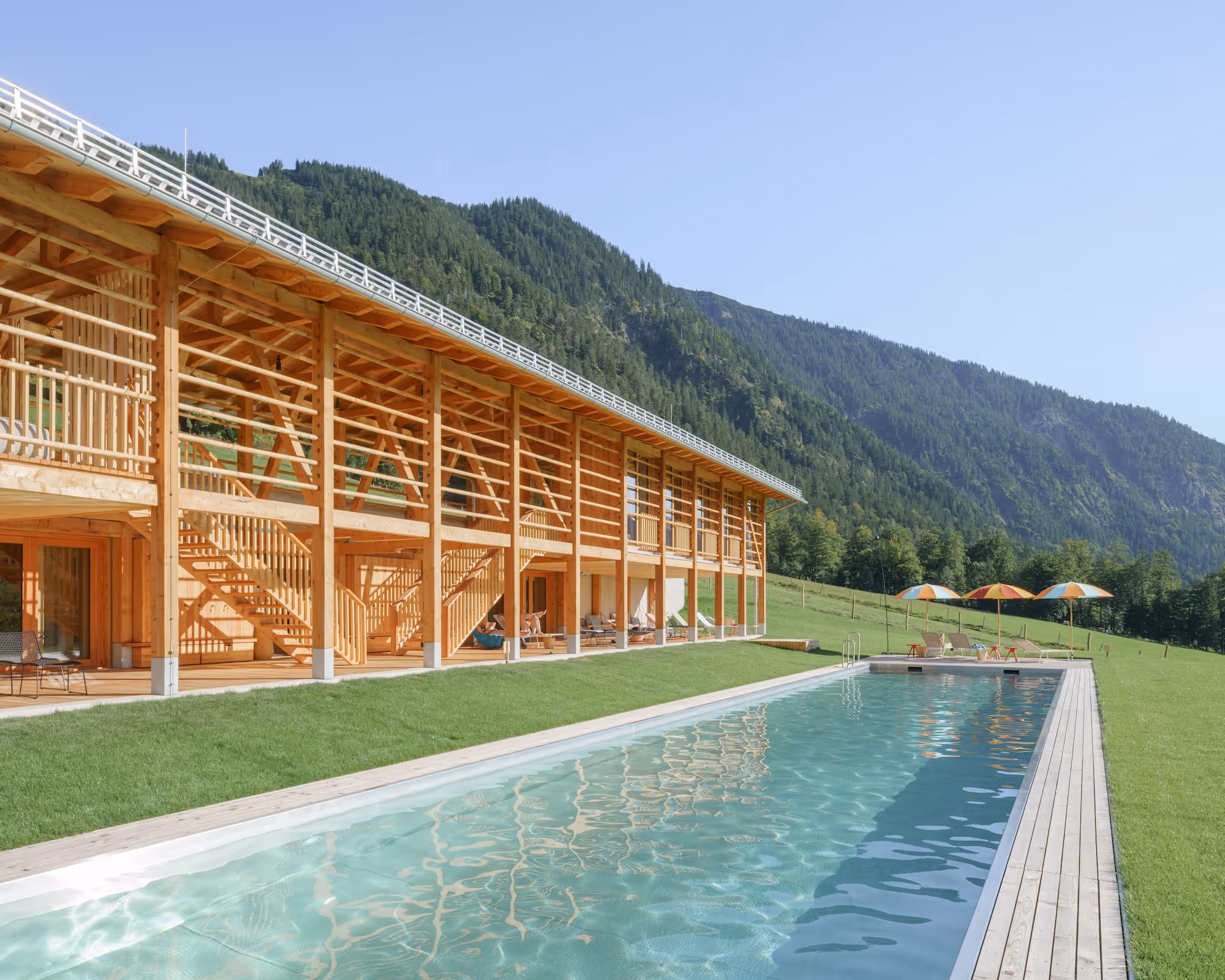 Modern wooden building with a long outdoor swimming pool and lounge chairs under umbrellas, set against green mountains and clear sky.