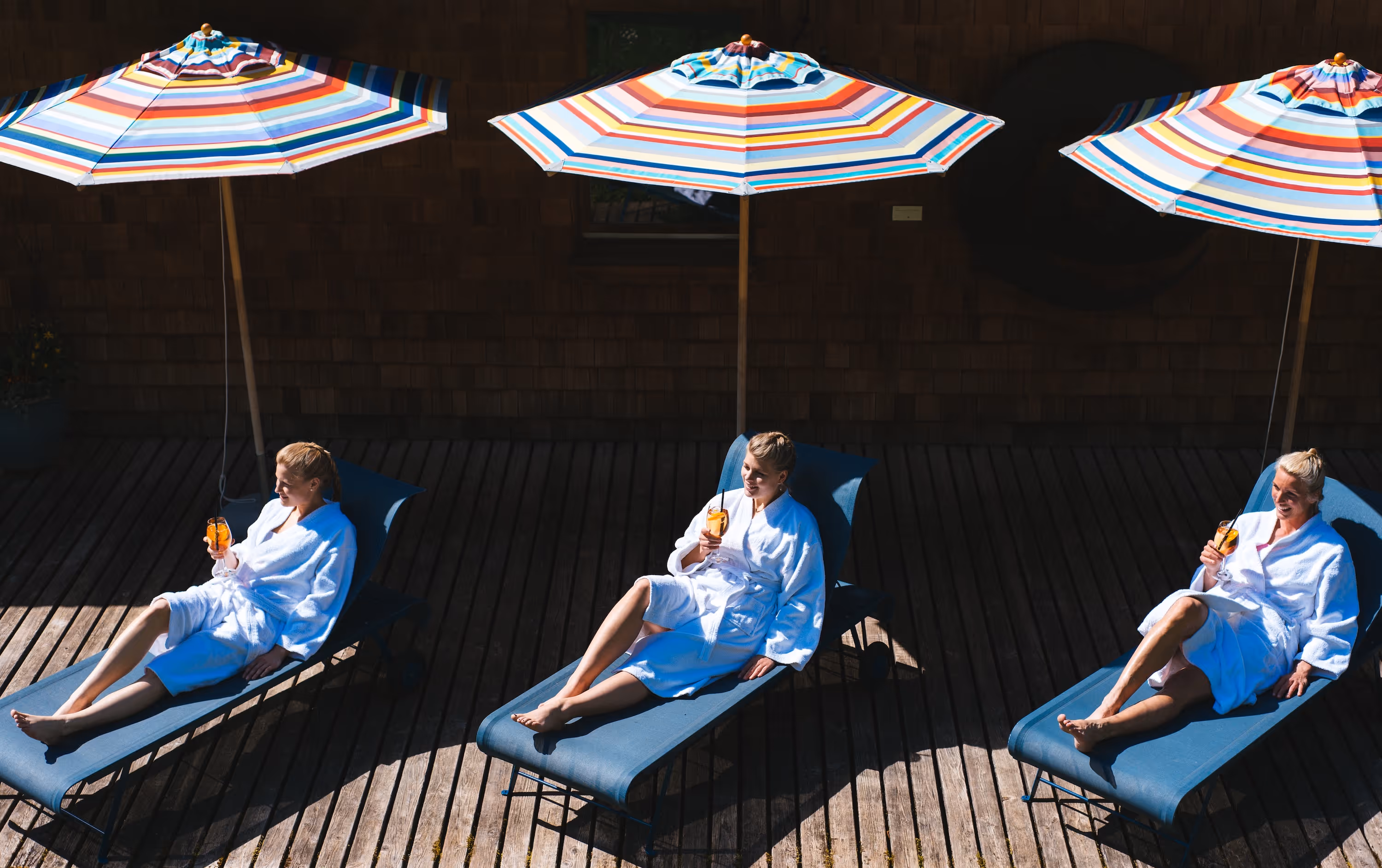 Three women in white bathrobes relaxing on blue lounge chairs under colorful striped umbrellas, each holding an orange drink.