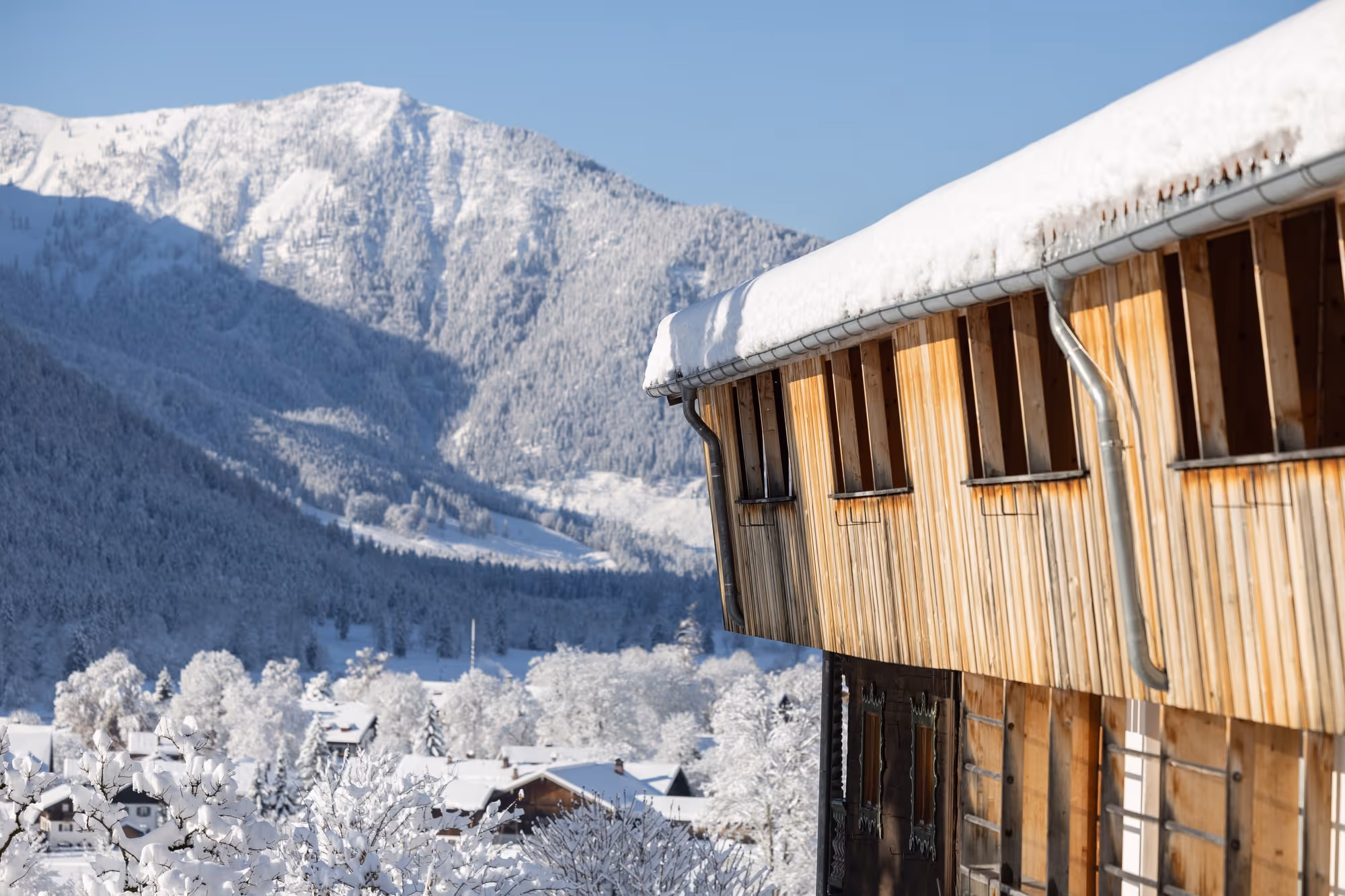 Snow-covered wooden chalet overlooking a snowy village and forested mountain under clear blue sky.