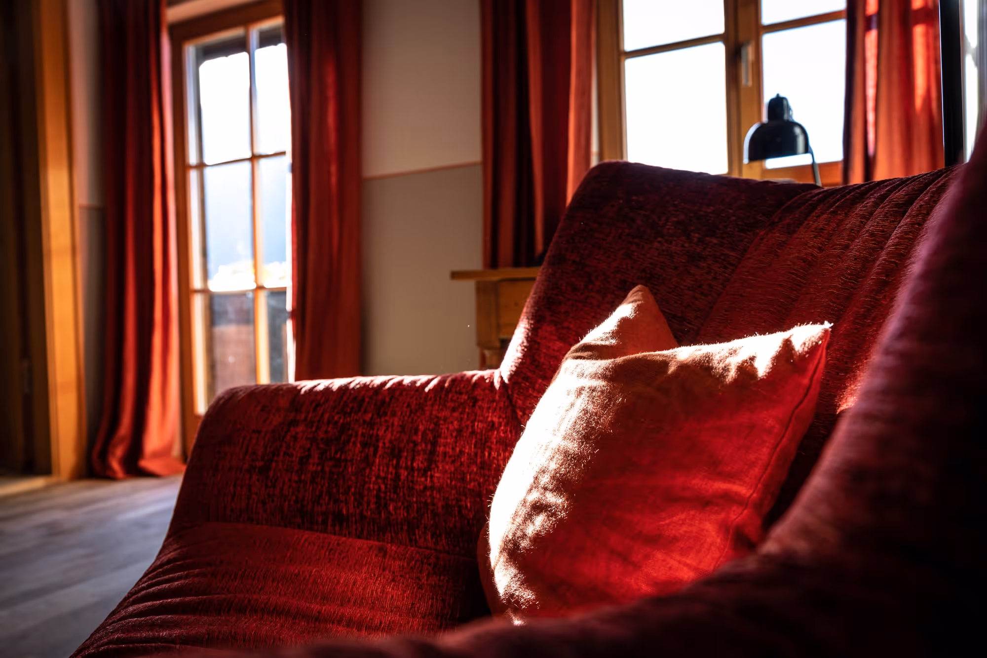 Sunlit red couch with a textured red pillow in a cozy room with red curtains and wooden framed windows.