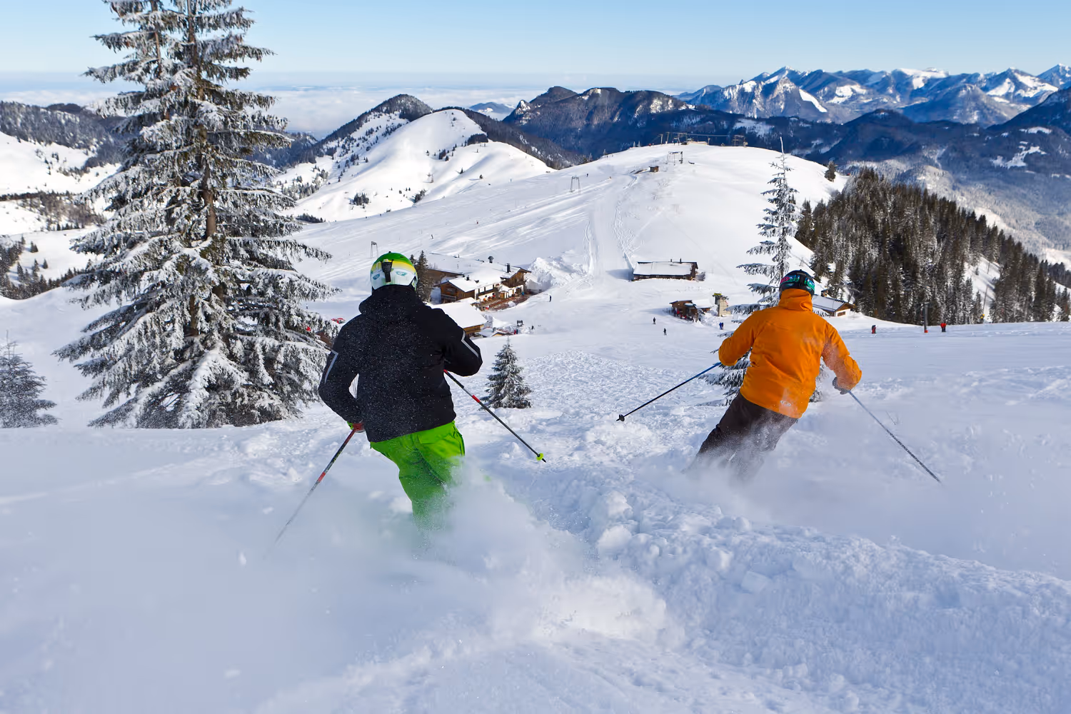 Zwei Skifahrer in bunten Jacken fahren durch frischen Pulverschnee auf einem schneebedeckten Berg mit Blick auf eine alpine Hütte und Bergkette.