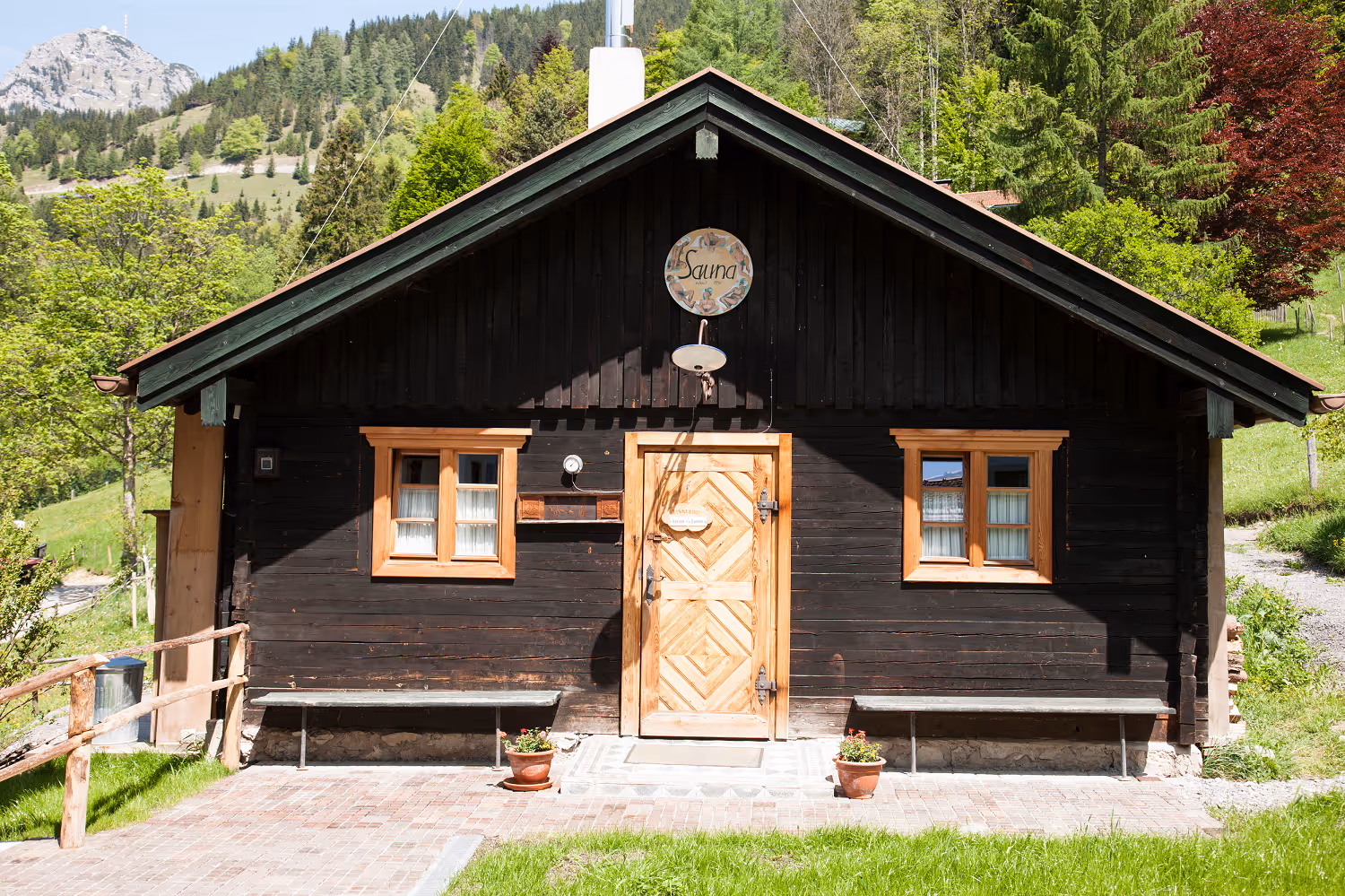 Rustic wooden sauna cabin with a chevron-patterned door, two windows, benches, and potted plants in a green mountainous landscape.