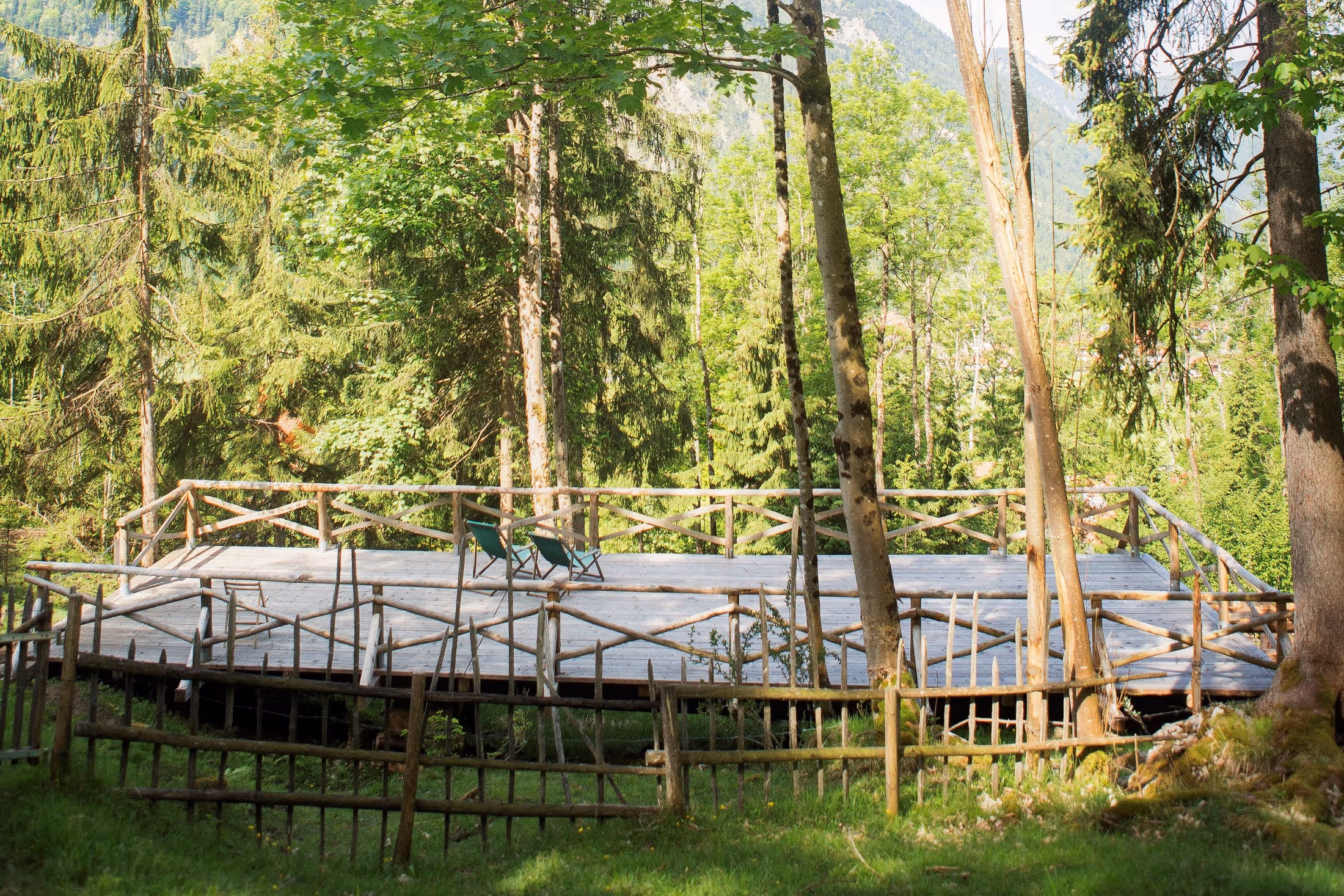 Wooden sun deck with two green lounge chairs surrounded by trees and mountainous forest scenery.