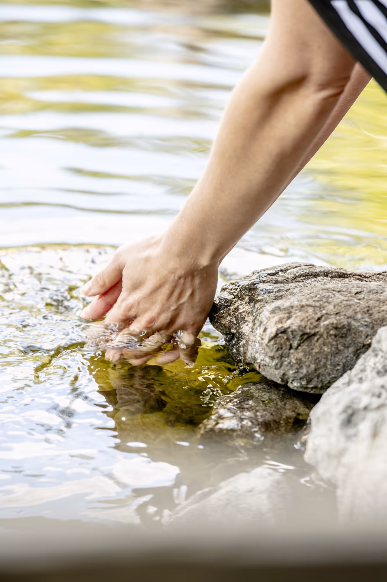 A hand touching clear water near a rocky edge, creating ripples.