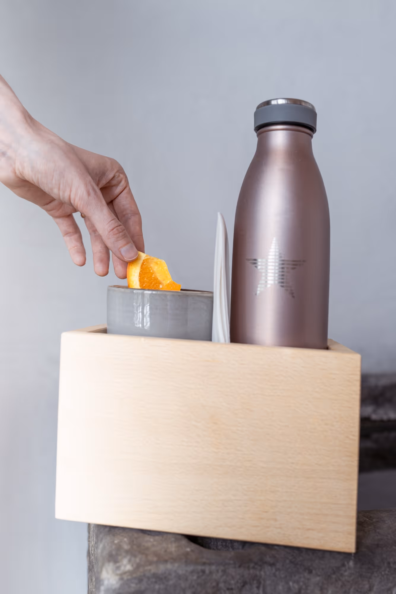 Hand placing an orange wedge into a gray cup next to a metallic water bottle with a star design, all in a wooden holder.