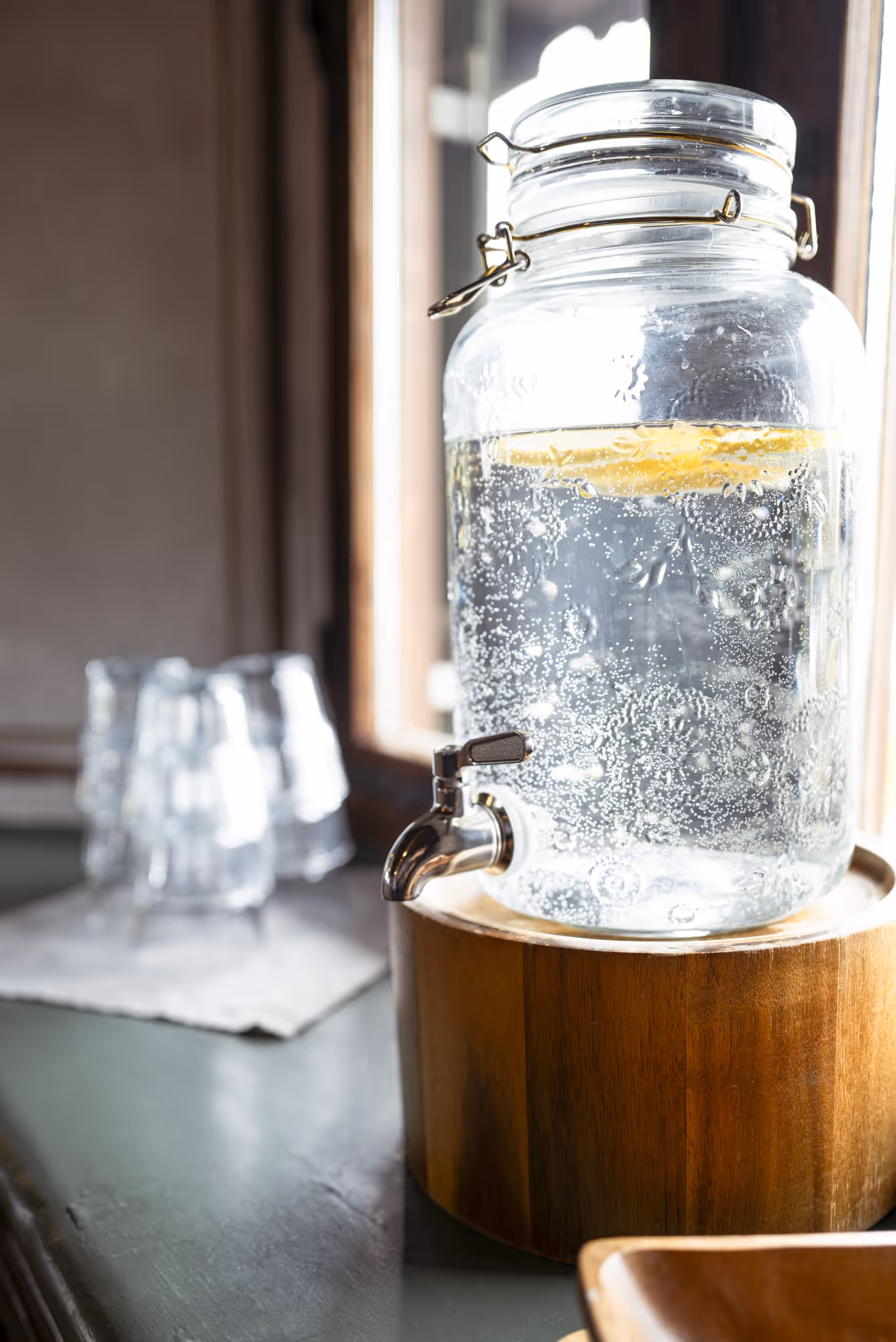 Glass water dispenser with lemon slices and bubbles on a wooden stand near a window, with empty glasses in the background.