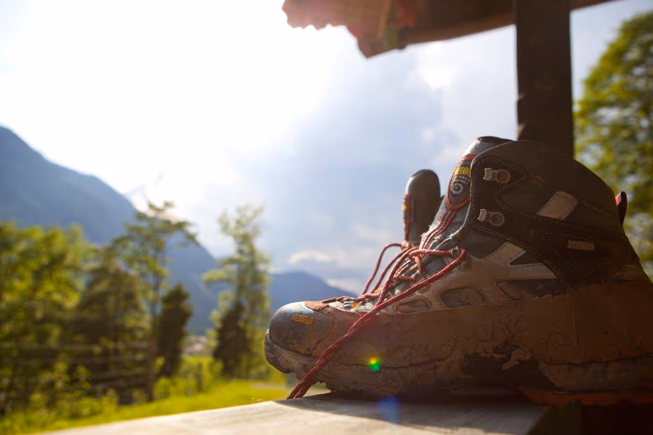 Schmutzige Wanderschuhe mit roten Schnürsenkeln auf einem Holztisch vor bergiger Sommerlandschaft.
