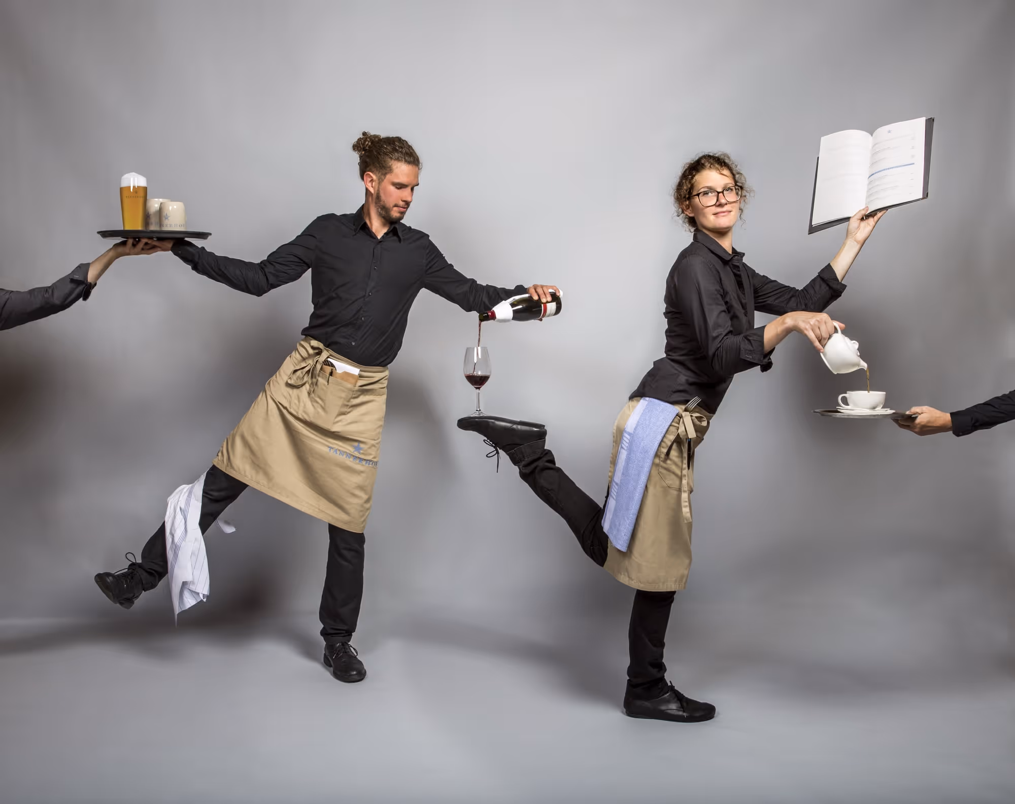 Zwei Kellner in Bewegung, der eine gießt Wein, die andere Tee und hält ein Buch hoch.