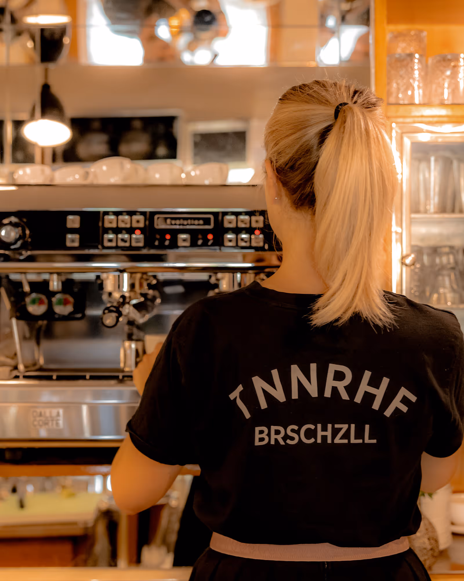 Kellnerin mit Pferdeschwanz steht vor Siebträgermaschine und bereitet Kaffee zu.