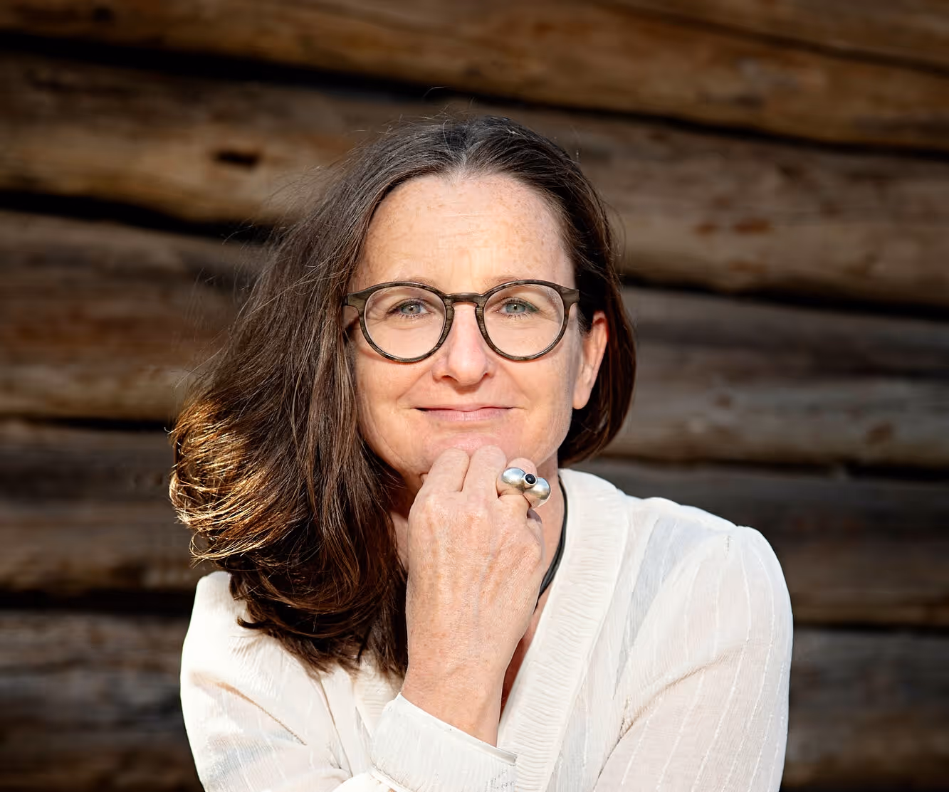 Frau mit braunen Haaren, trägt Brille und weißen Pullover, vor Holzwand, lächelt.