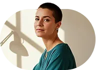 Young woman with short hair wearing a teal top, looking at the camera indoors near a window.