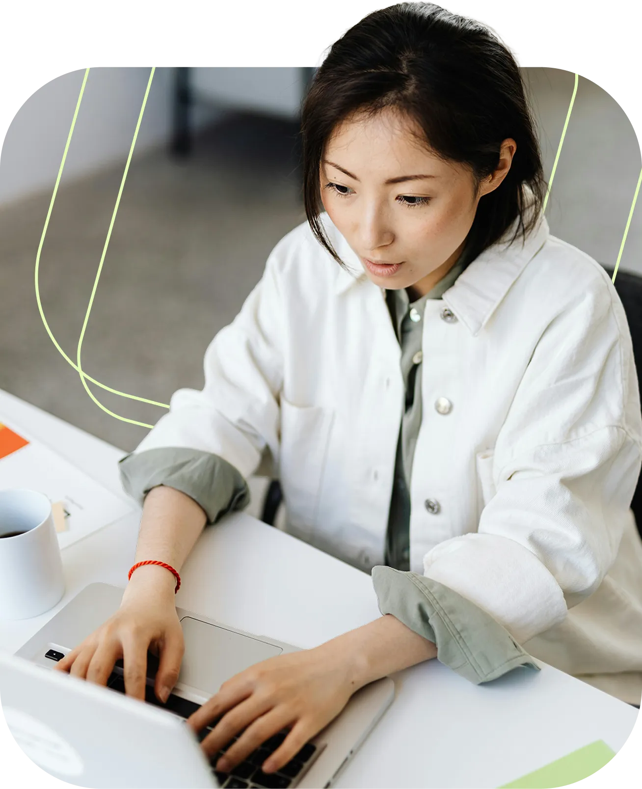 Woman in white jacket focused on typing on a silver laptop at a white desk with a coffee cup.
