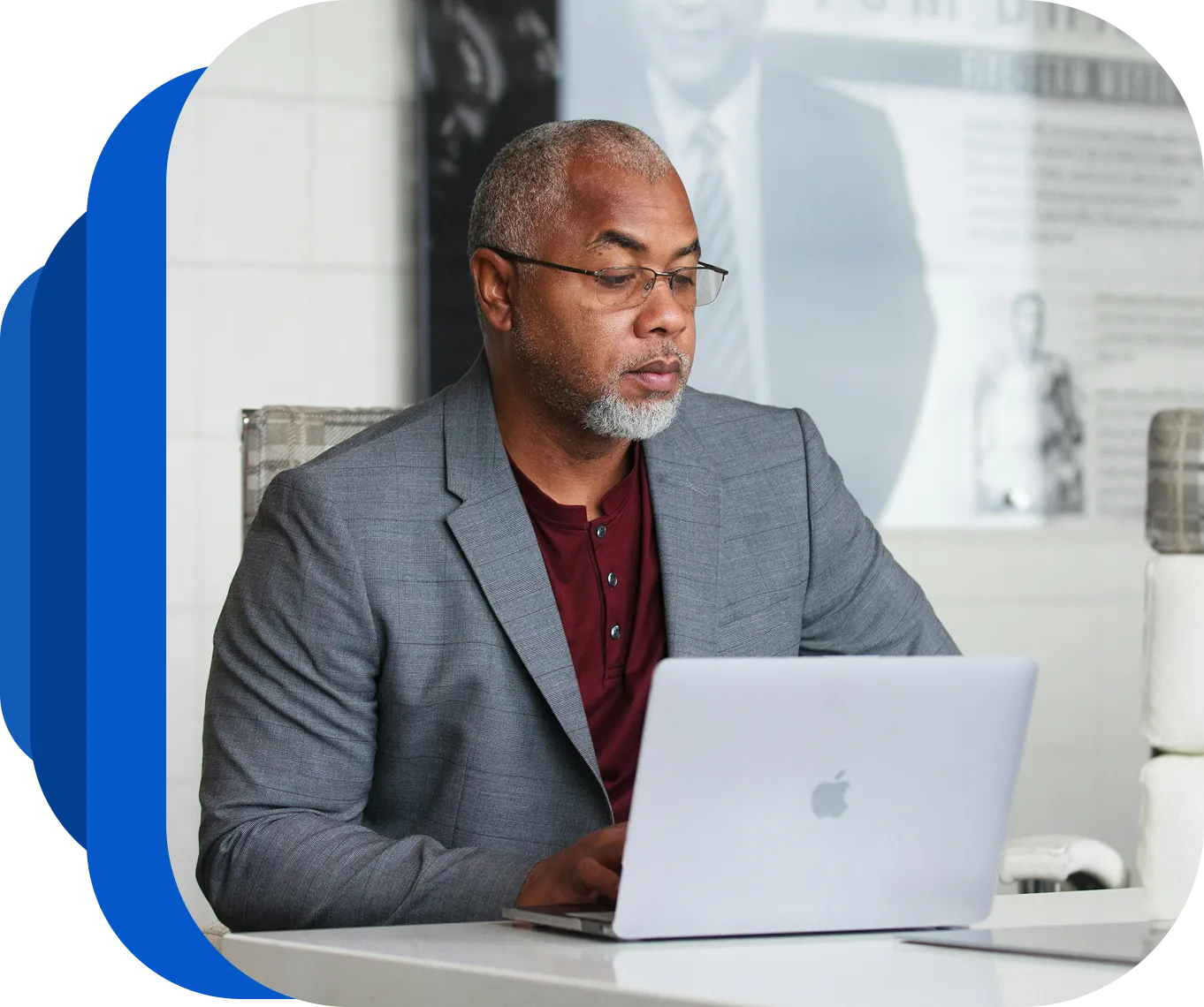 Man in glasses and gray blazer working on a silver Apple laptop at a white desk.
