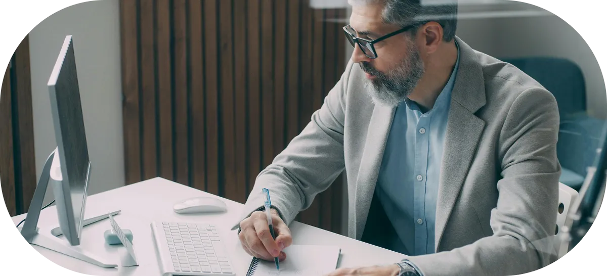Man with glasses and beard writing notes at a desk with a computer and keyboard.