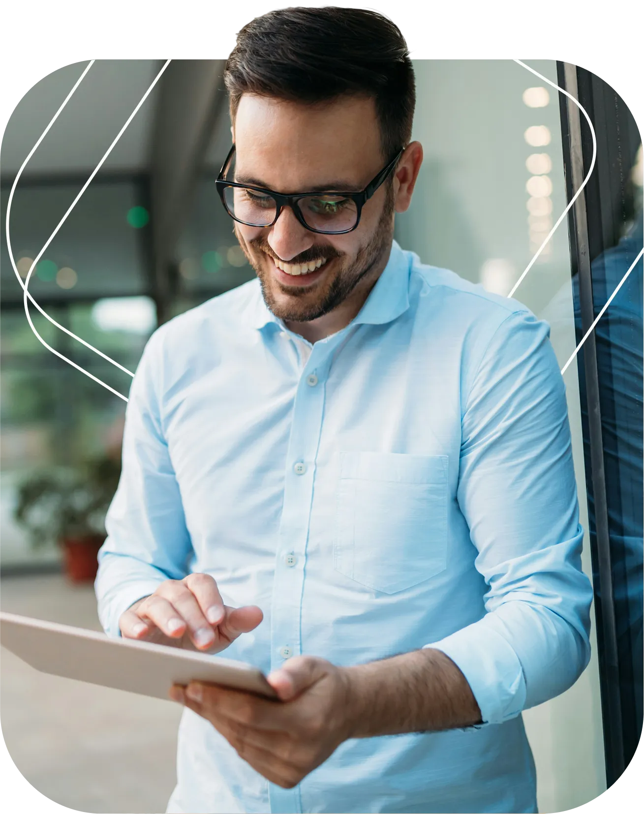 Smiling man with glasses using a tablet while standing indoors near a glass wall.