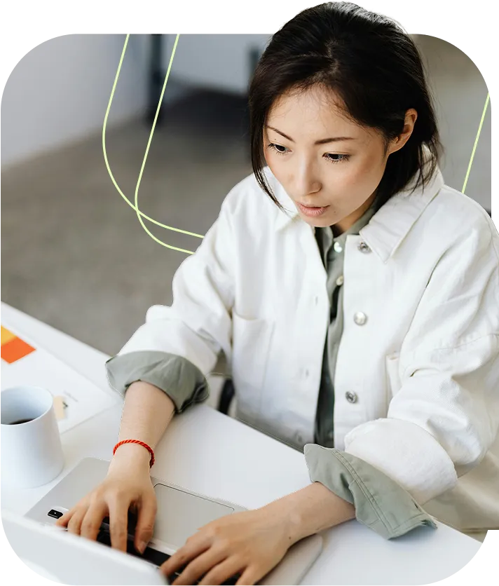 Woman wearing white jacket typing on a laptop with a cup of coffee on the desk.