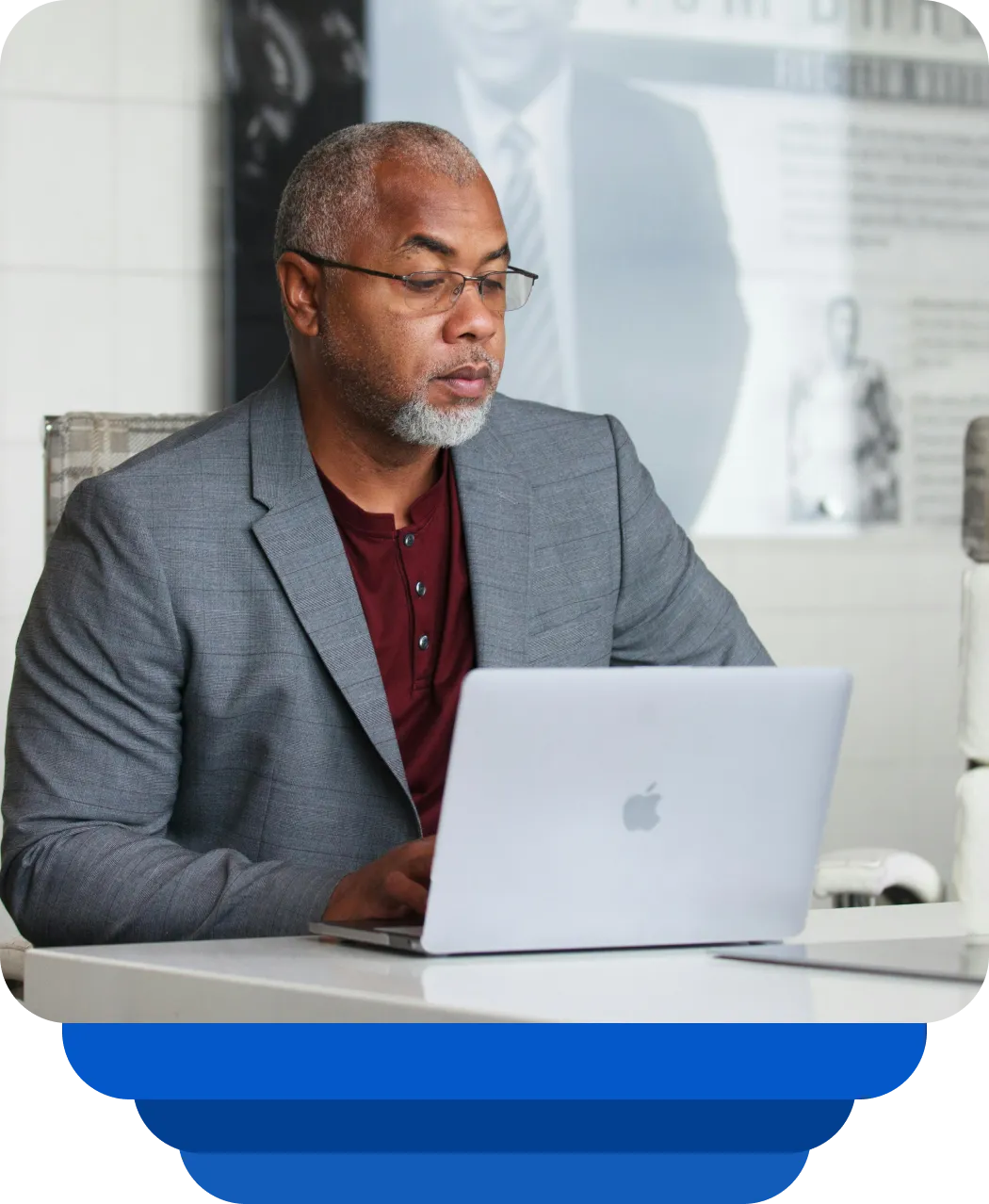 Older man wearing glasses and gray blazer working on a MacBook laptop at a white desk.