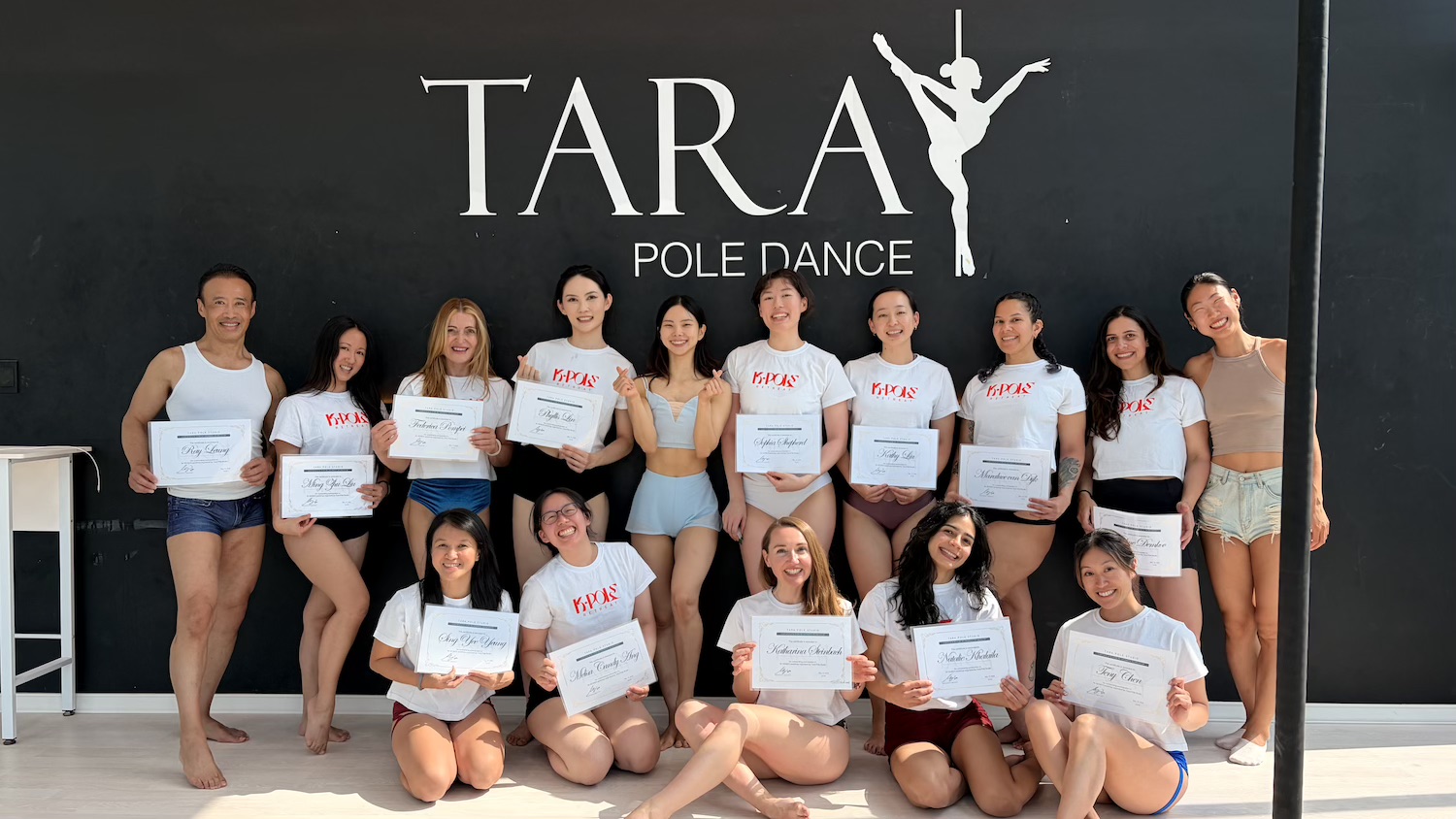 Smiling K-Pole dance students showing certificates in front of a black wall with a TARA POLE DANCE logo.