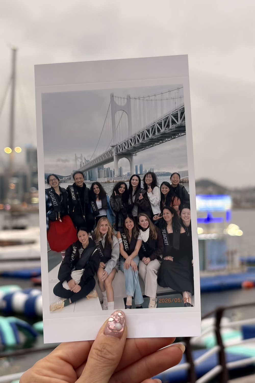 Hand holds a polaroid photo of a K-Pole retreat participants posing on a boat under Gwangalli suspension bridge in Busan, Korea.