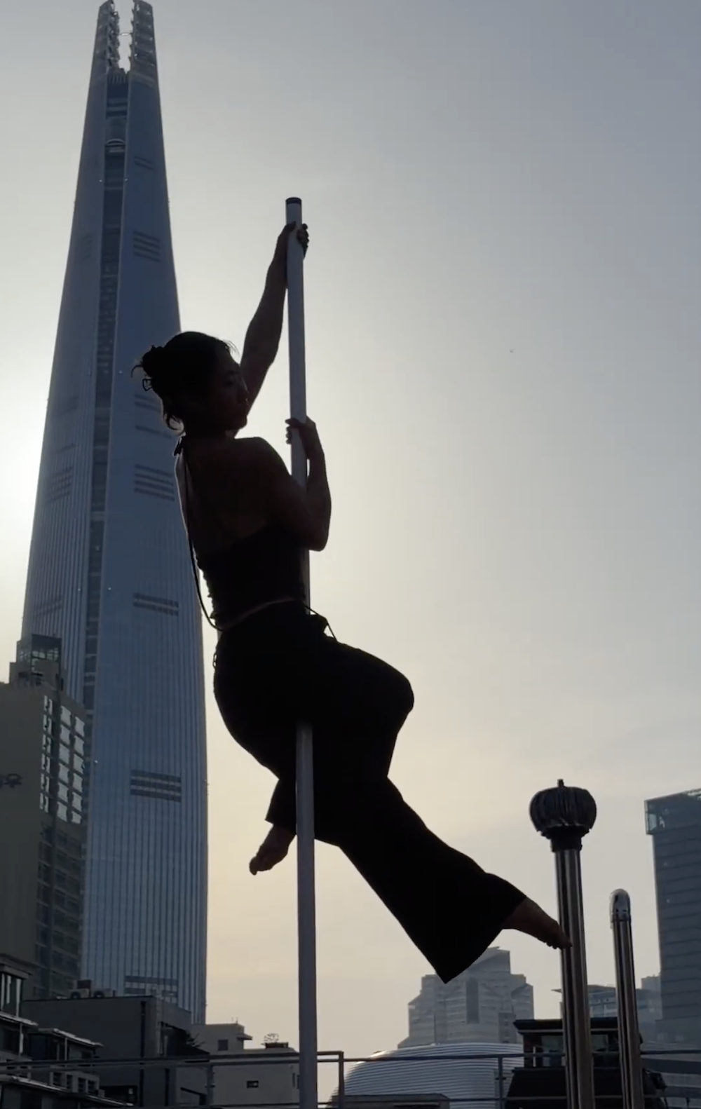 Haeri doing a pole dance trick on a rooftop with Jamsil tower and the Seoul City skyline in the background.