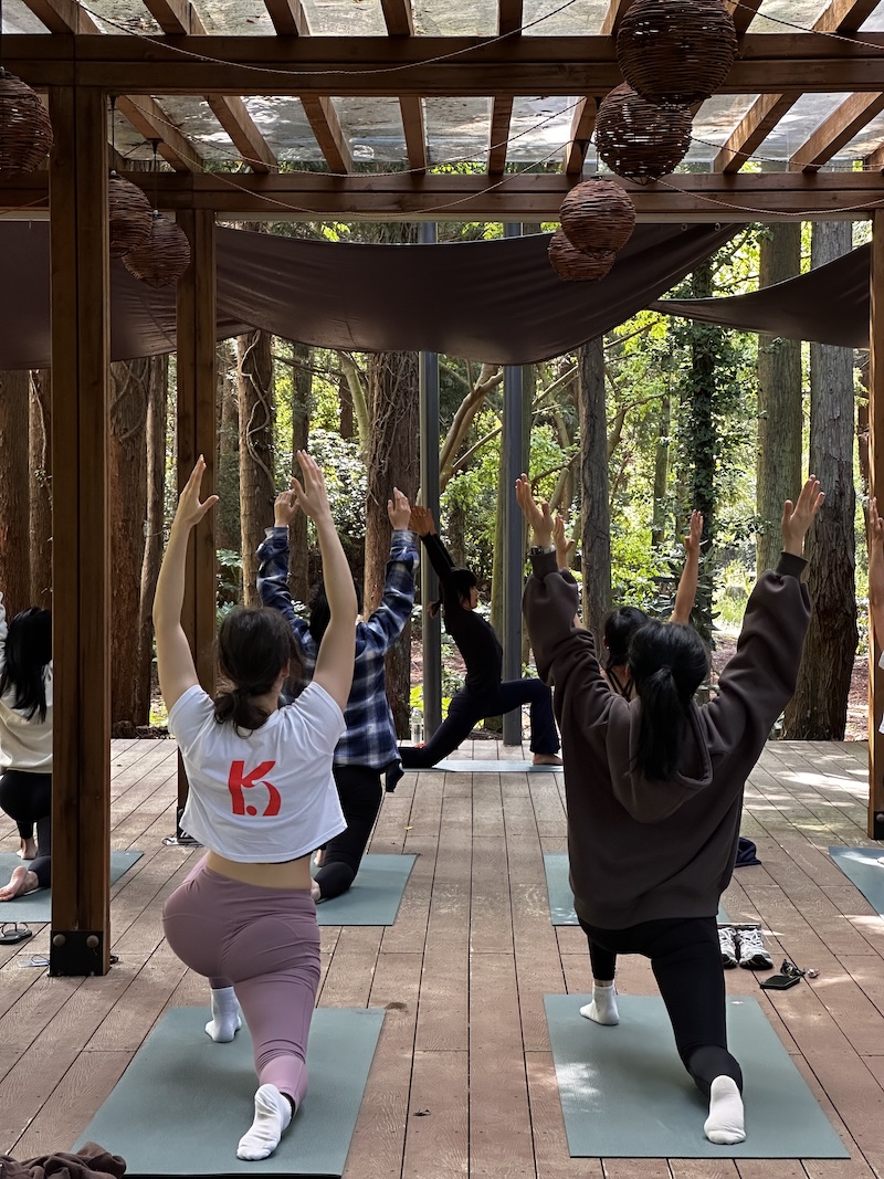 K-Pole family practicing yoga on mats under wooden pergola surrounded by trees in outdoor setting.