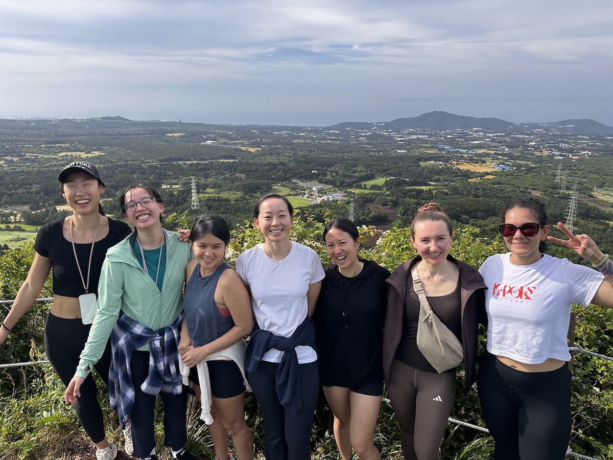 K-Pole participants group photo after climbing a hill on Jeju Island. The background shows more hills and a great view of the surrounding area.
