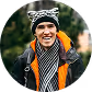 Smiling woman wearing a striped scarf, black jacket, gray beanie, and backpack outdoors.