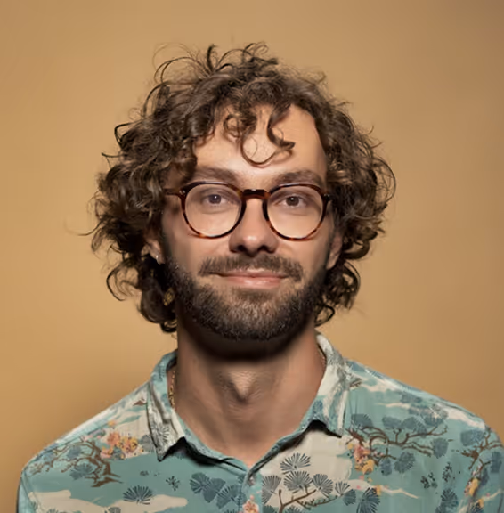 Man with curly hair, a beard, glasses, and a floral patterned shirt against a beige background.