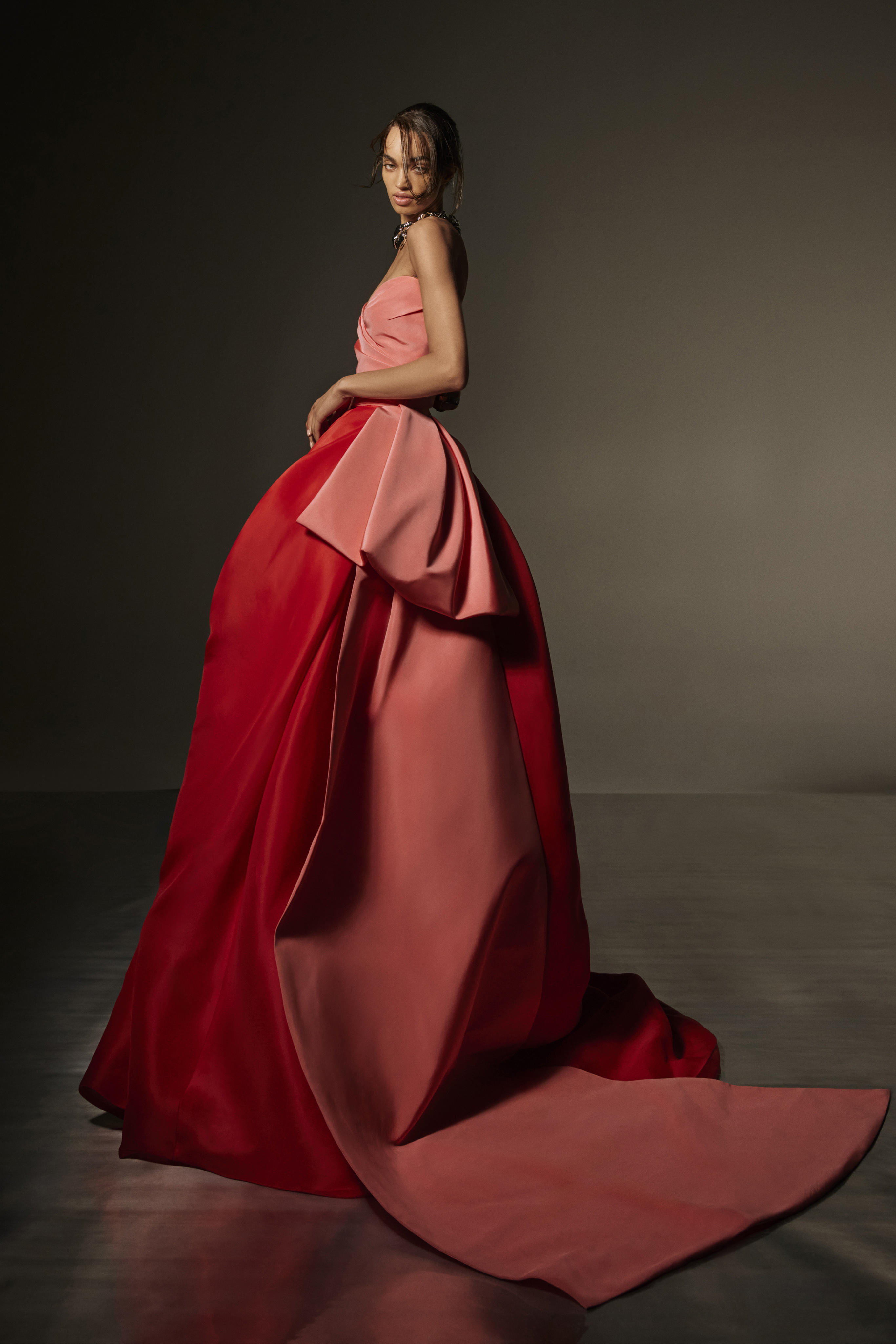 Woman in red and pink strapless ball gown with flowing train posing against a dark background.
