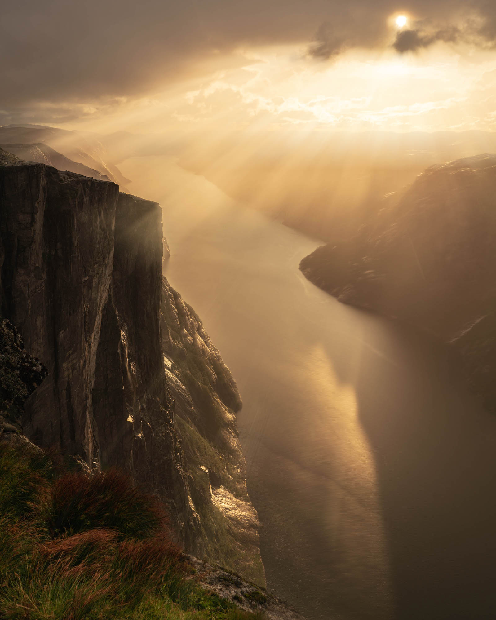 Norway — towering cliffs above a sunlit Norwegian fjord, golden light piercing dramatic storm clouds