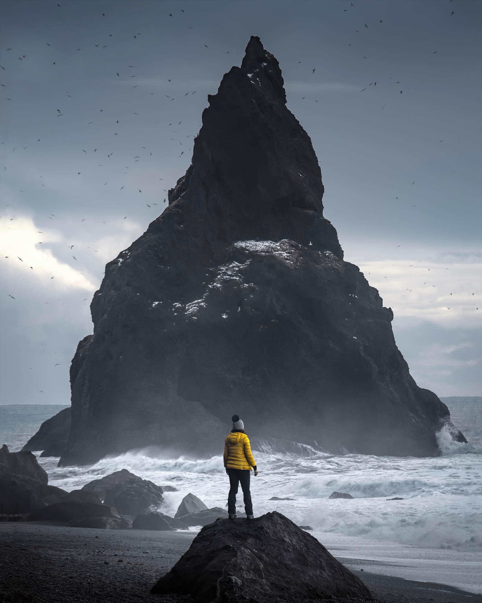 Iceland — a lone traveller standing before a towering basalt sea stack on Reynisfjara’s black sand beach