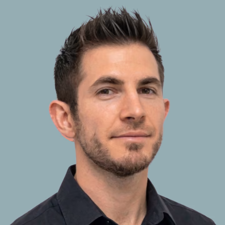 Portrait of a man with short spiked hair and trimmed beard wearing a black shirt against light blue background.