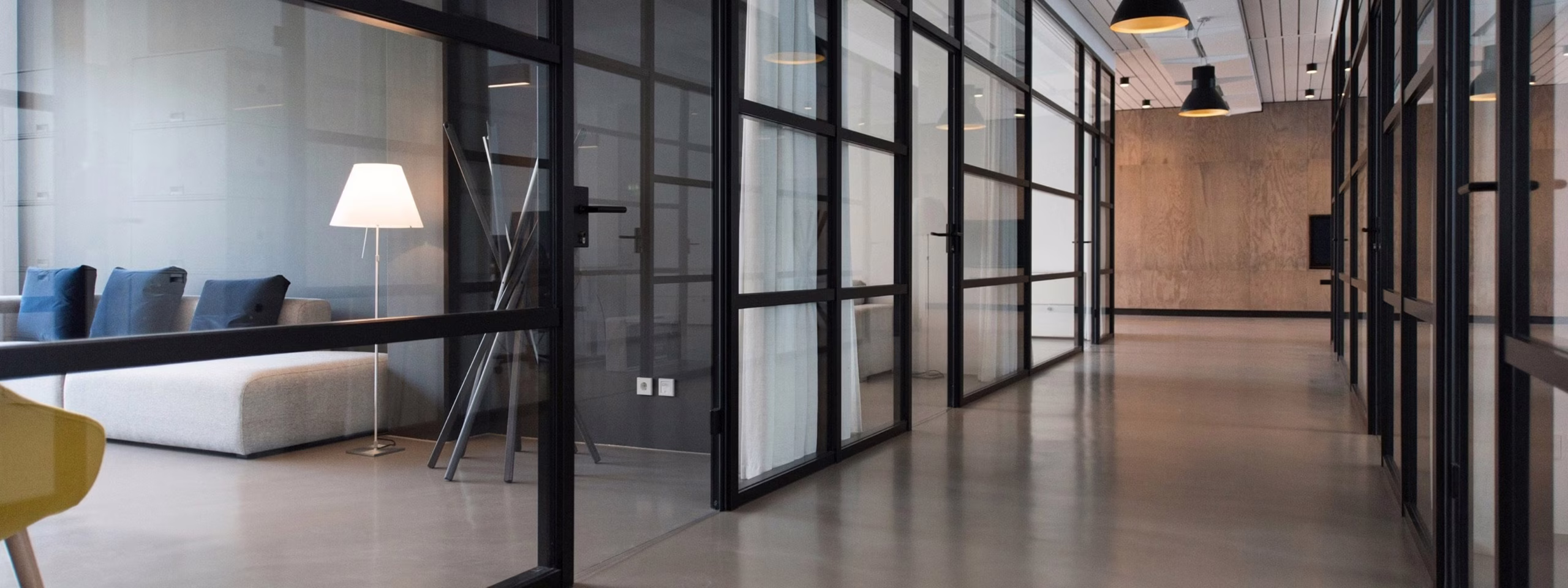 Modern office hallway with glass walls, gray sofa, floor lamp, and ceiling pendant lights.