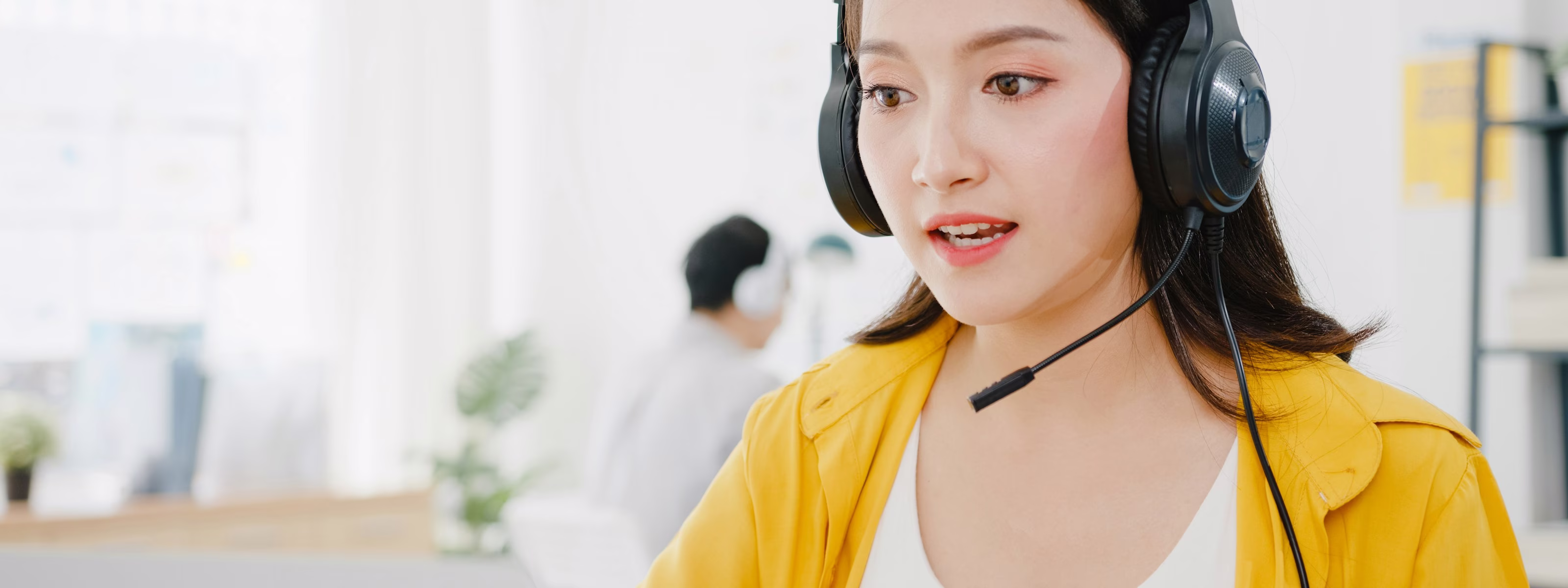 Woman in yellow jacket wearing headset with microphone, speaking and working in a bright office.