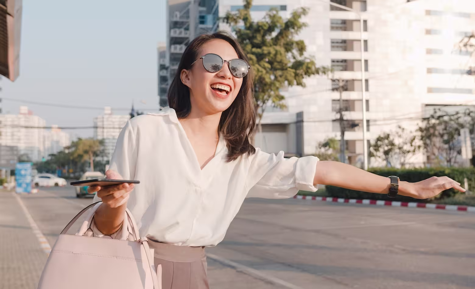 Smiling woman with sunglasses holding phone and handbag, hailing a ride on city street.