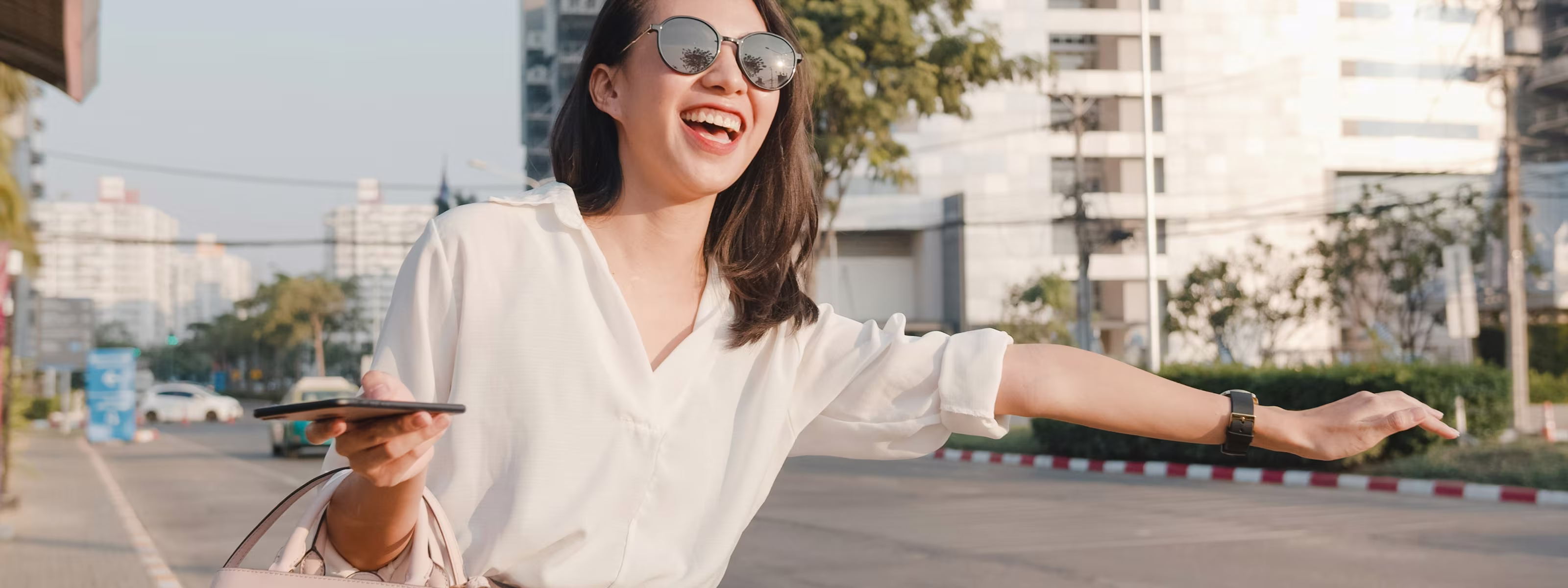 Smiling woman in sunglasses holding phone, raising arm to hail a ride on city street.