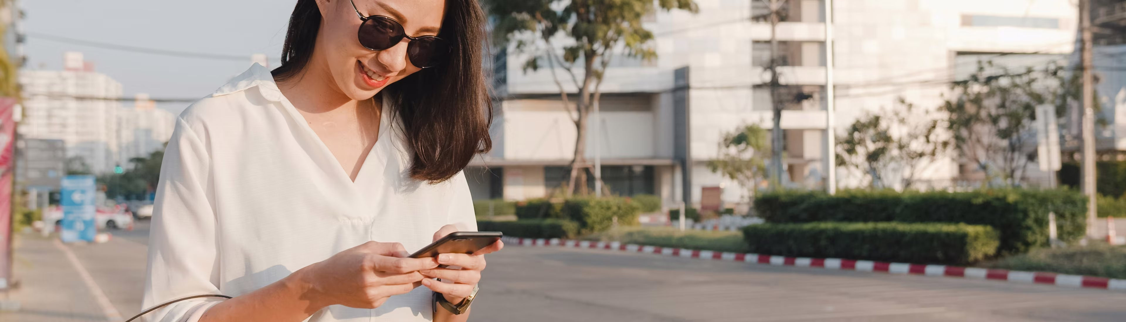 Smiling woman in sunglasses looking at her phone outdoors on a sunny city street.