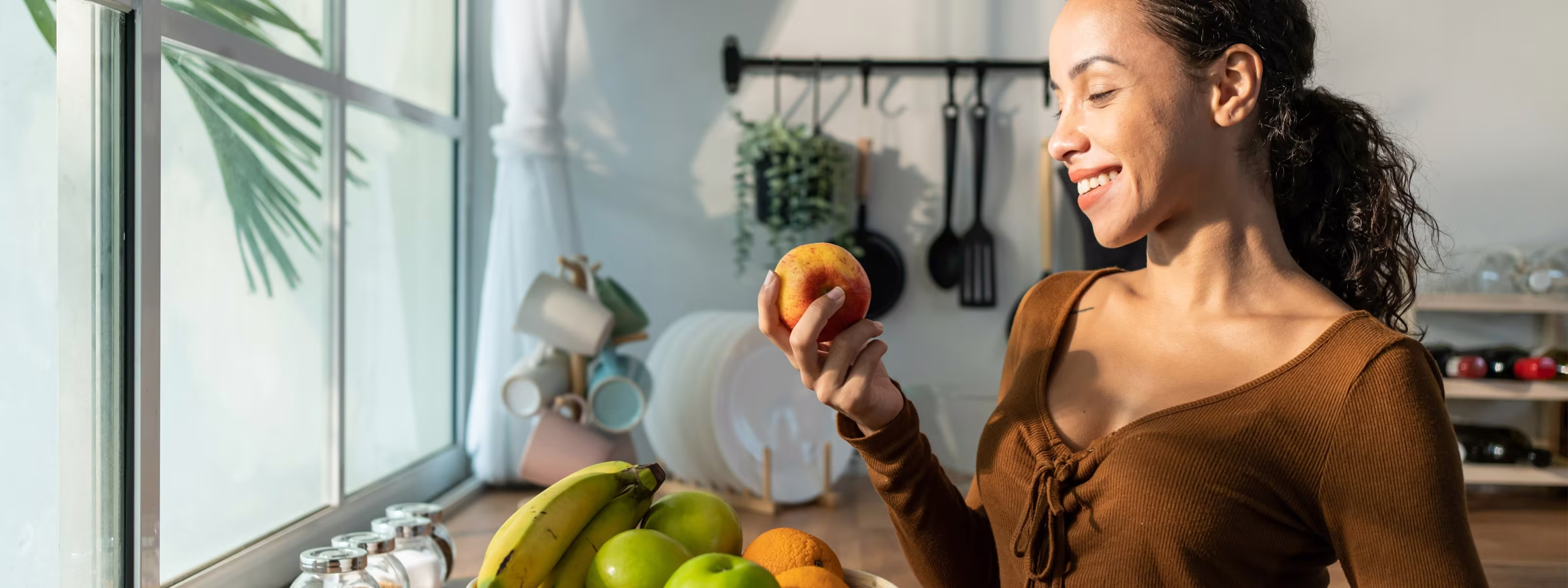 Woman smiling and holding an apple in a kitchen with bananas, apples, and oranges on the counter.