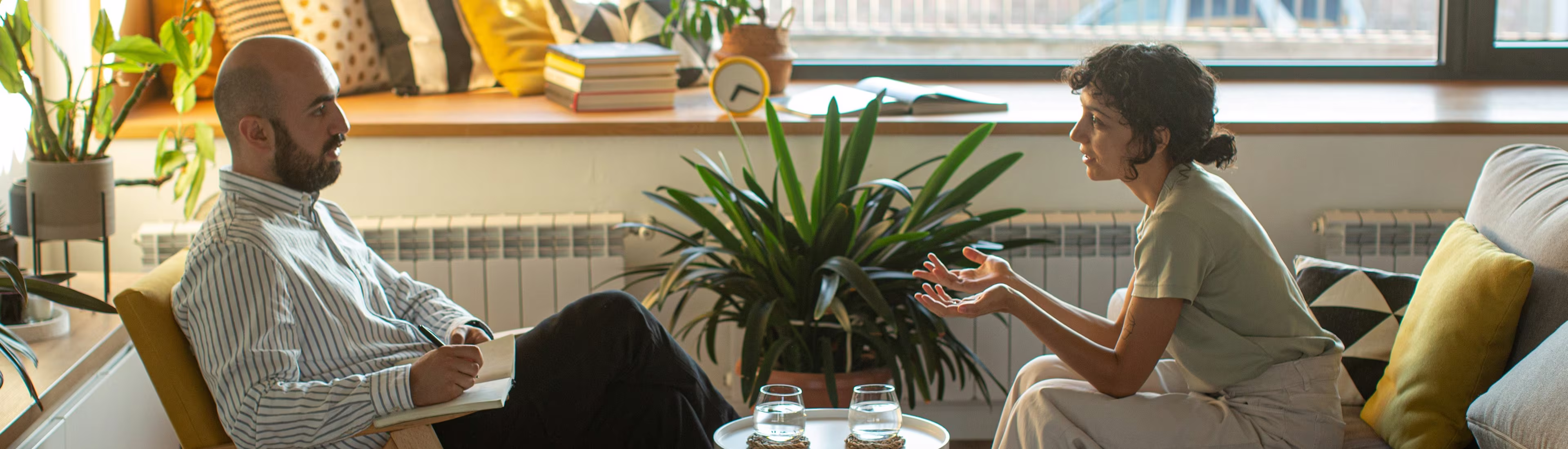 Man with notebook listens while woman talks in a cozy, plant-filled living room setting.