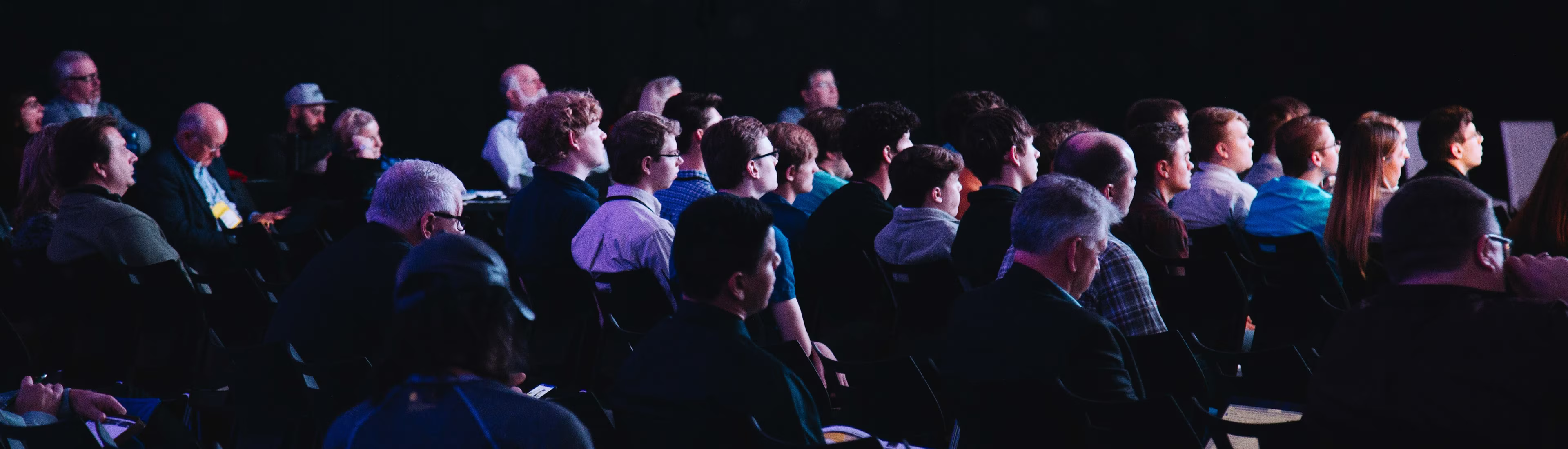 Audience of men and women attentively watching a presentation in a dark room.