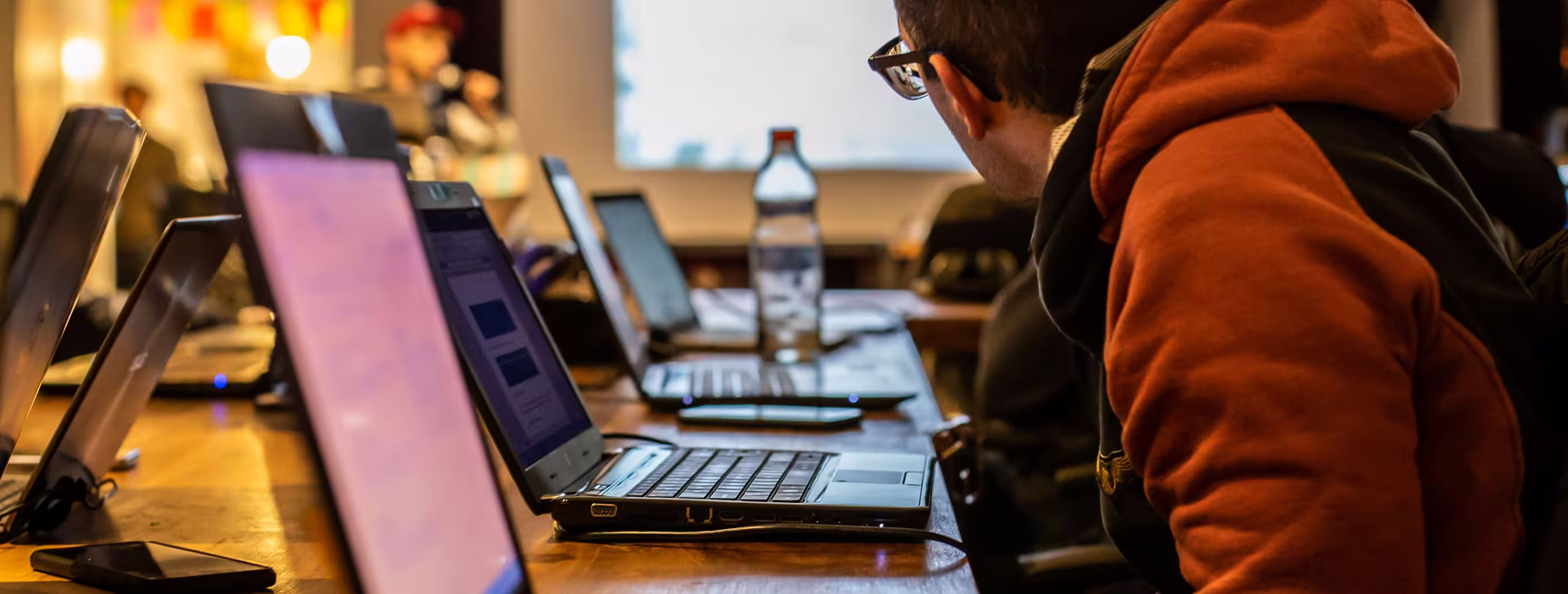 Person in an orange hoodie works on a laptop at a busy shared table, surrounded by computers and a water bottle in a warm, focused workspace.