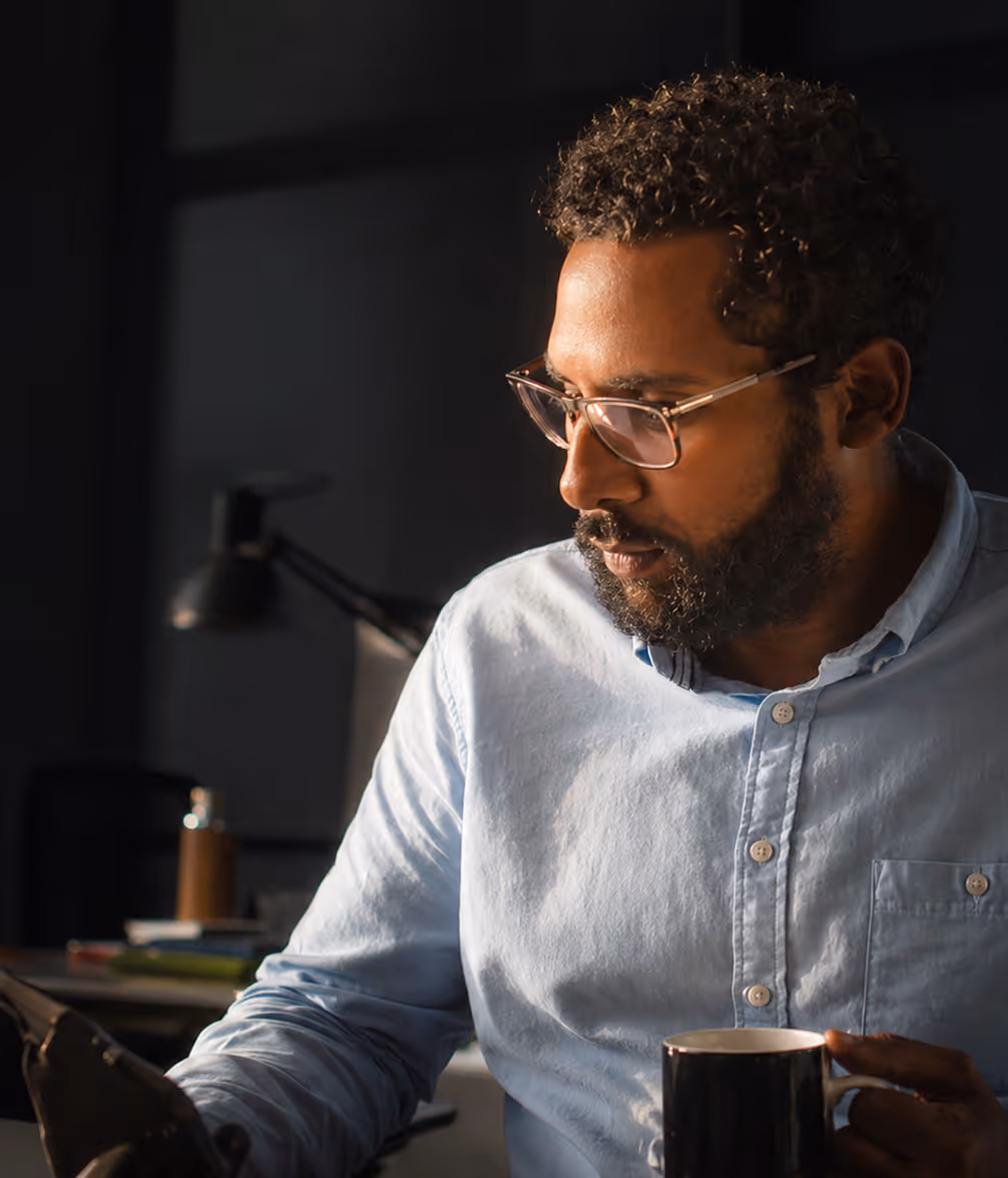 Man with curly hair and glasses wearing a blue shirt, holding a mug and looking at a tablet in a dimly lit room.